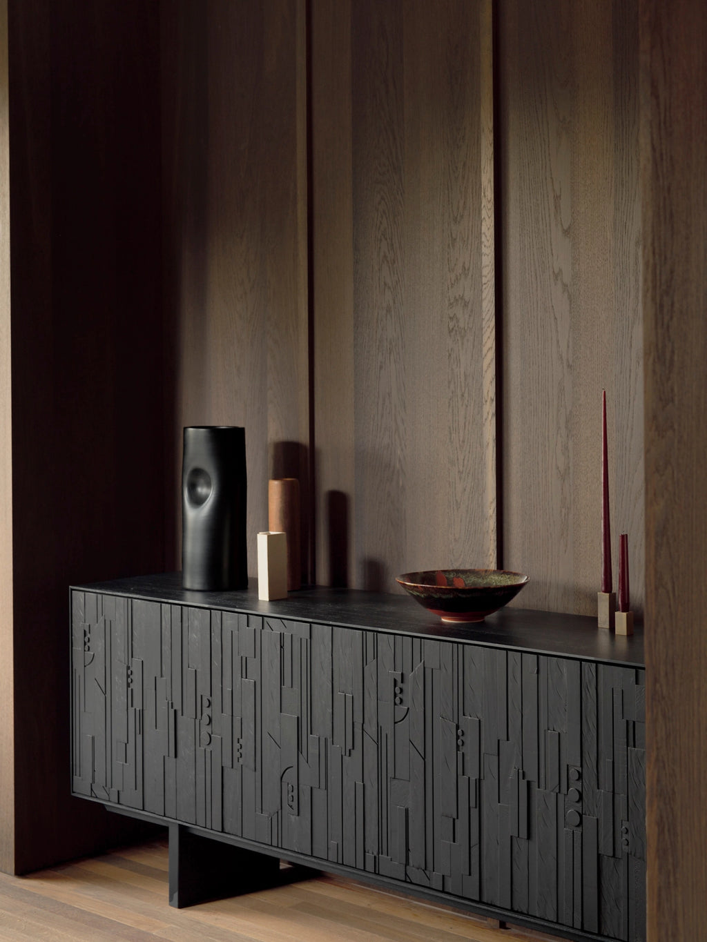 Ethnicraft Mosaic Sideboard in Teak Off-Black, a sophisticated dark wood cabinet featuring intricate carved patterns, elegantly placed in a room with rich wooden walls and decorative items.