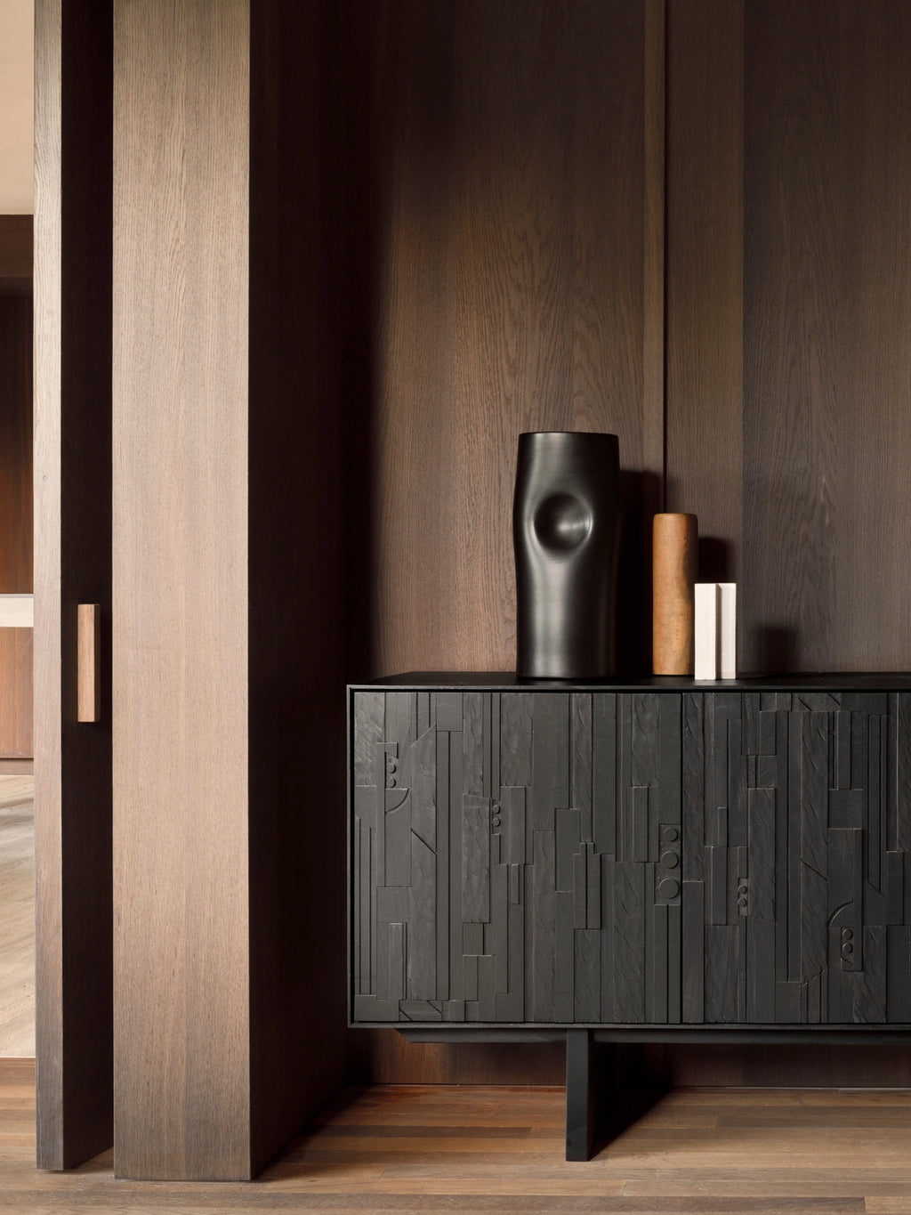 Ethnicraft Mosaic Sideboard in Teak Off-Black, a stylish dark wood cabinet with carved patterns, positioned in a modern living space with warm wood paneling and decorative accents.