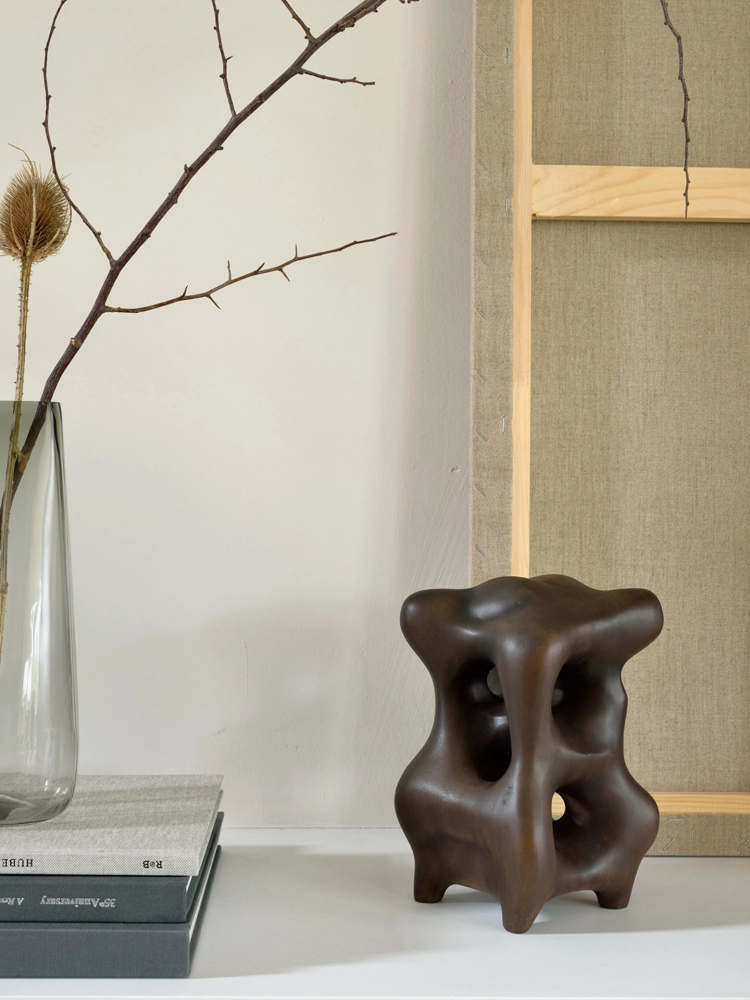 Ethnicraft Natural Organic Sculpture, a dark brown mahogany piece with an organic, abstract form, displayed as a decorative accent on a white shelf next to a vase with dried branches and stacked books.