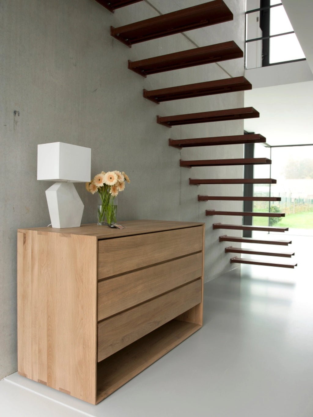 Ethnicraft Nordic Dresser, a minimalist light oak wood dresser with three spacious drawers and an open bottom shelf, styled in a contemporary hallway. A white lamp and vase of flowers adorn the top, complementing the modern concrete wall and floating staircase.