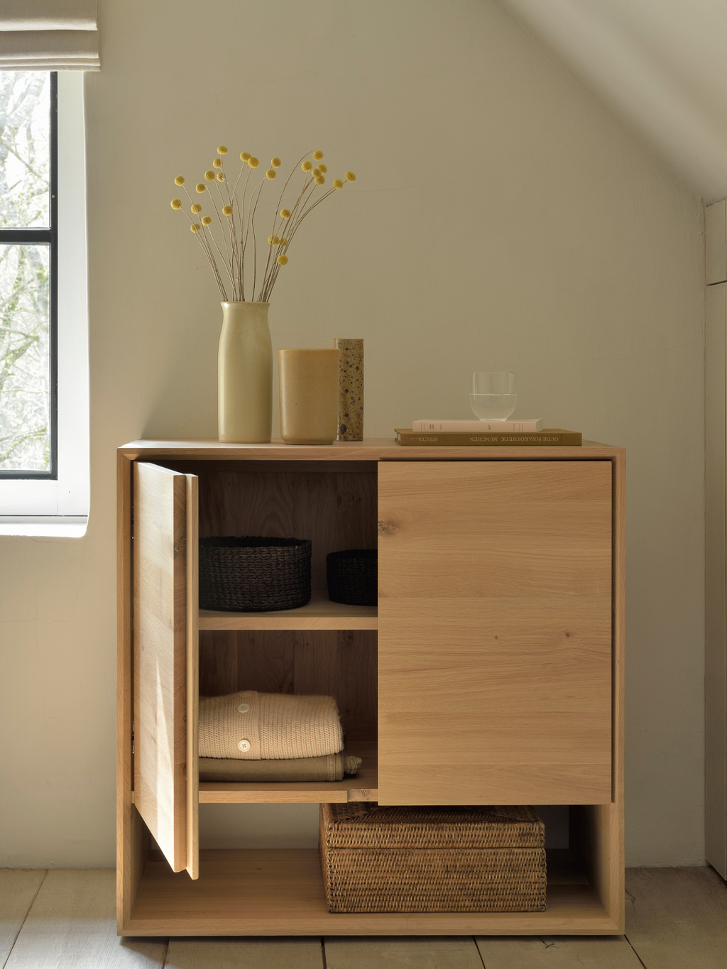 Ethnicraft Nordic Sideboard in natural oak, a classic and functional storage unit with two doors, one open revealing shelves with folded clothes and baskets, styled with a vase of dried flowers in a bright room.