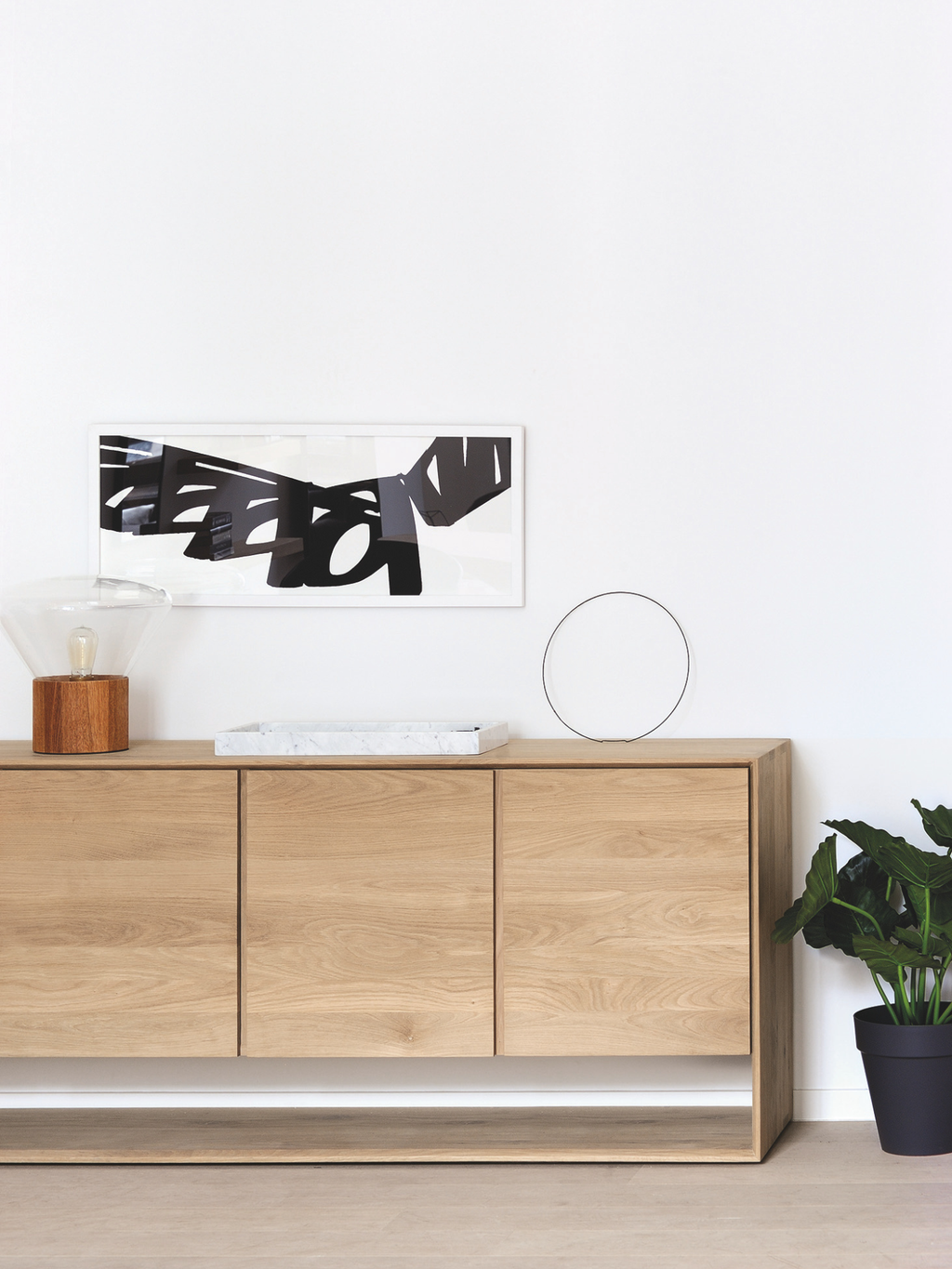 Ethnicraft Nordic Sideboard in a modern living room setting, featuring its light oak wood finish, styled with a contemporary lamp, framed art, and a green plant, against a white wall.
