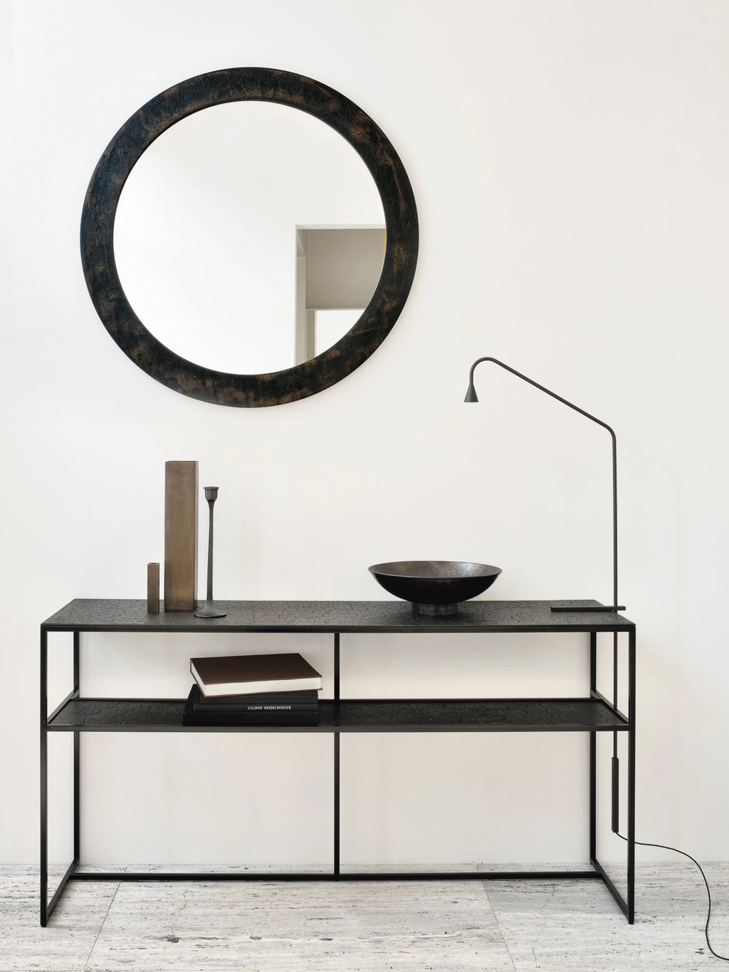Ethnicraft Pentagon Console in Whisky finish, elegantly styled in a minimalist room with a large round mirror above. The dark, textured console with its sleek metal frame holds various decorative accents, creating a sophisticated look.