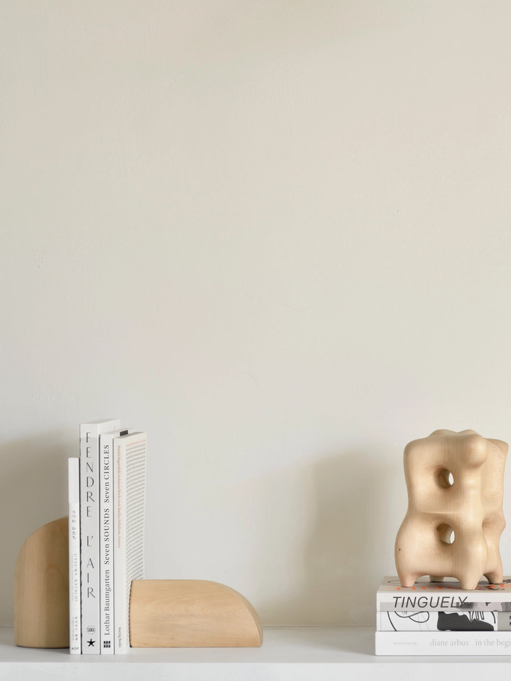 Ethnicraft PI Bookends, a pair of stylish sycamore wooden bookends holding a stack of books on a white shelf, complementing a modern, sculptural wooden decor piece in a minimalist room.