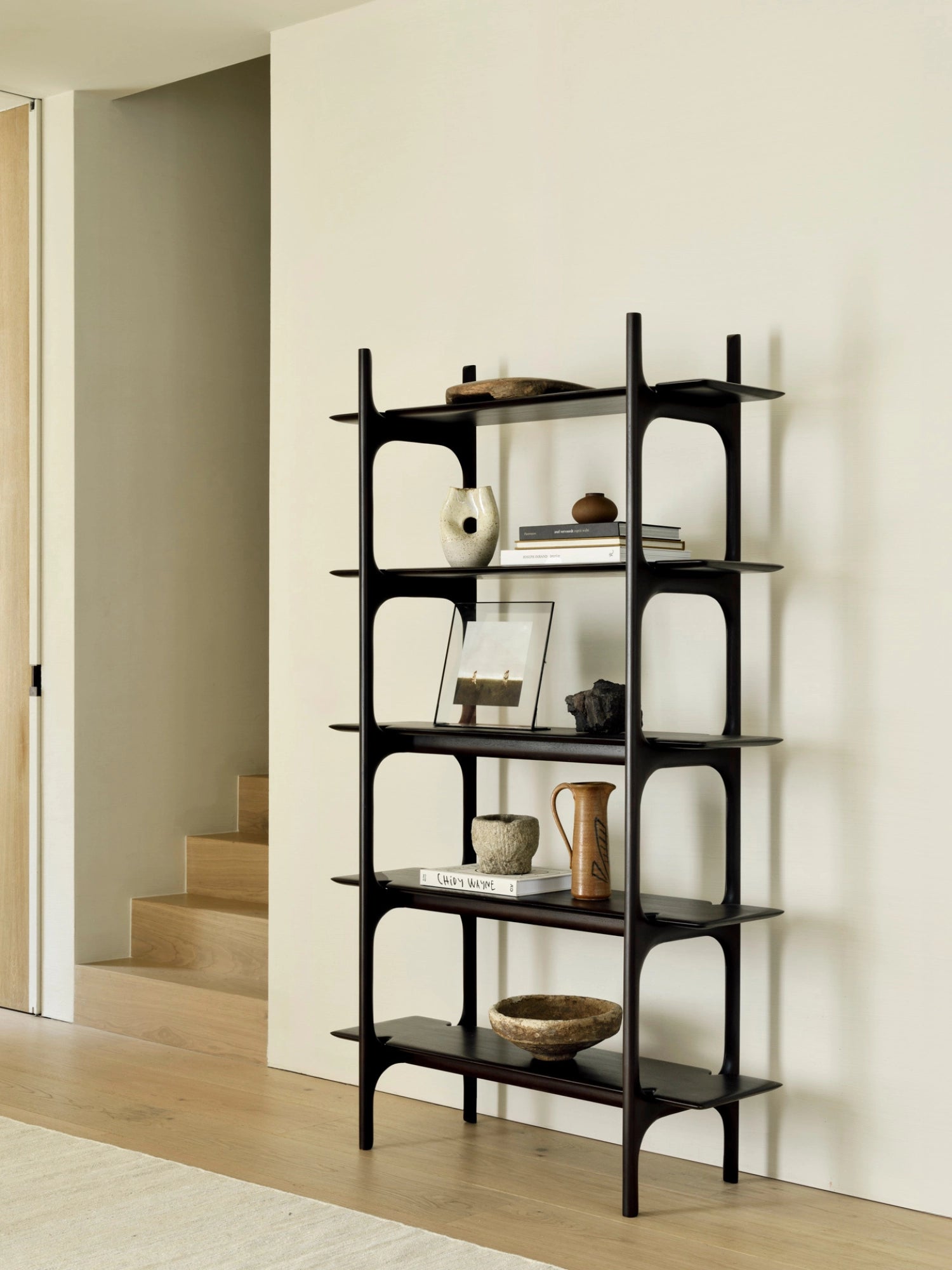 Ethnicraft PI Rack in rich mahogany, a dark brown, elegant shelving unit with five open shelves, adorned with various decorative objects, standing in a bright hallway.