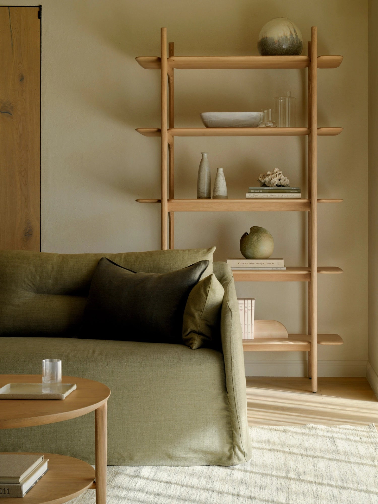 Ethnicraft PI Rack crafted from light oak wood, a tall, modern shelving unit with four open shelves, filled with decorative items, next to a green sofa in a living room.