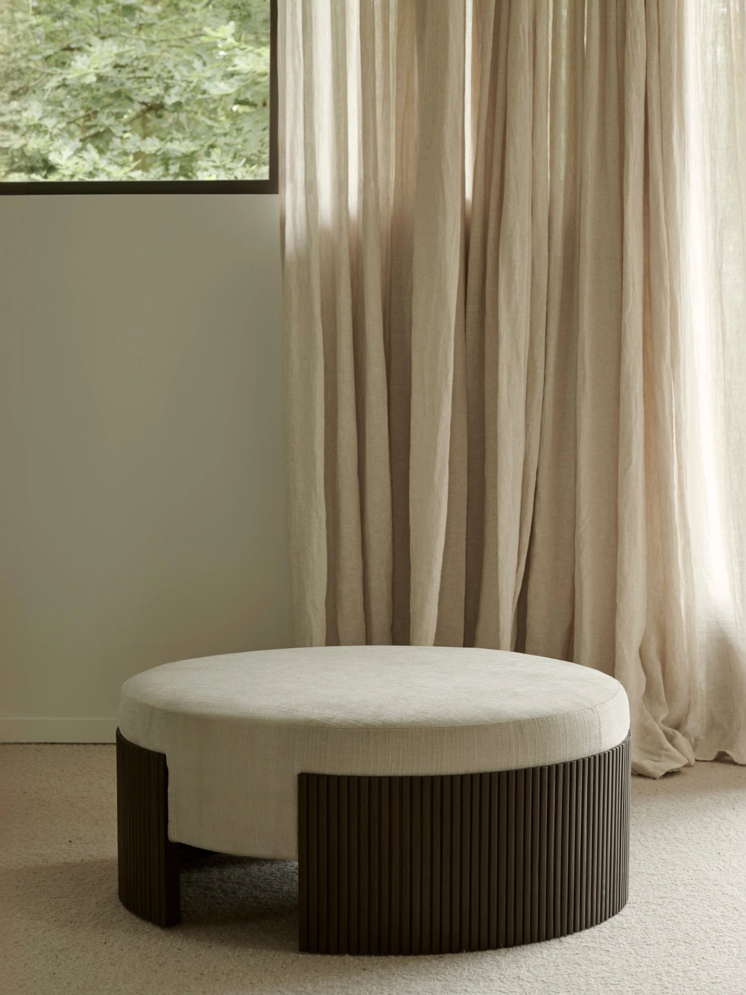 Ethnicraft Roller Max Pouf, 85cm wide, in mahogany, featured in a modern living space with light-colored curtains and a window, showcasing its elegant, round form and textured fabric.