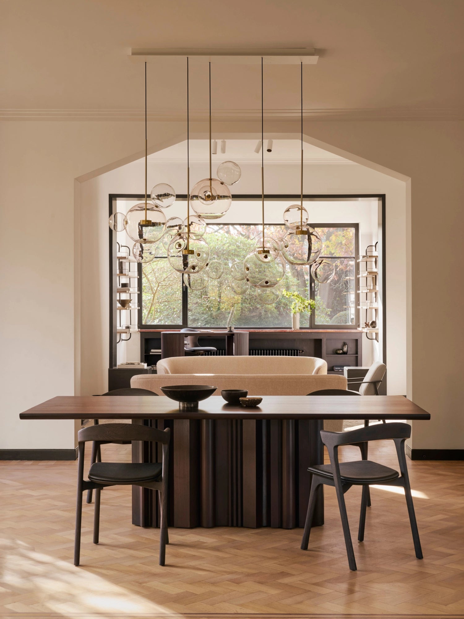 Ethnicraft Sequence Dining Table, a luxurious mahogany dark brown table with a fluted pedestal base, surrounded by dark wood chairs. The elegant dining room features a large window, a modern chandelier, and a comfortable sofa in the background, creating a refined atmosphere.