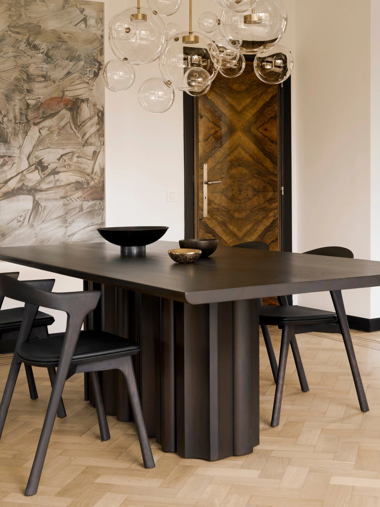 Ethnicraft Sequence Dining Table, a sophisticated mahogany dark brown table with a distinctive fluted base, paired with sleek black chairs. The setting features a large abstract painting and a unique wooden door, creating an artistic, luxurious dining space.