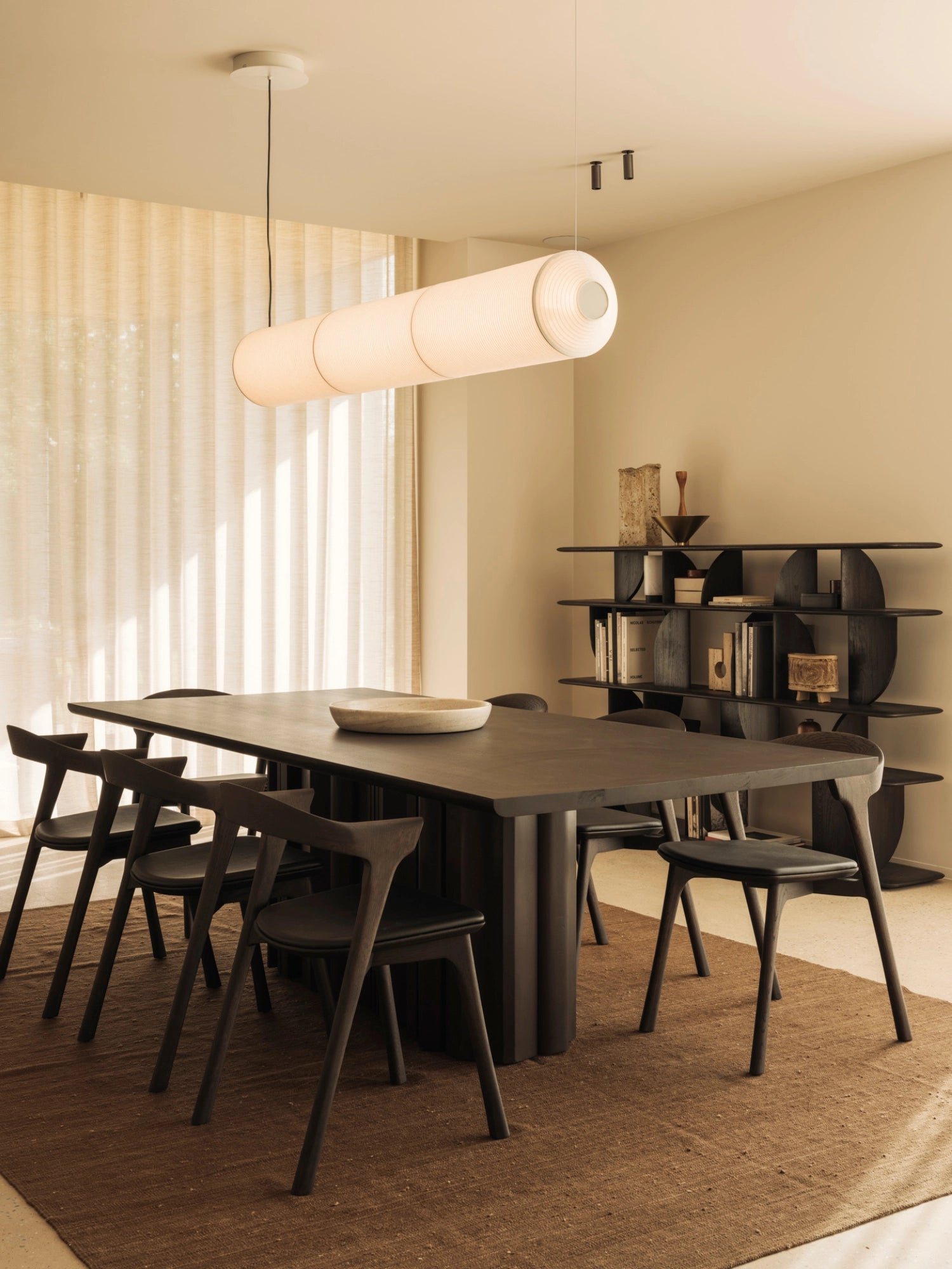 Ethnicraft Sequence Dining Table, a grand mahogany dark brown table with a fluted base, surrounded by matching dark wood chairs in a warm, inviting dining room. A long, cylindrical pendant light hangs above, enhancing the modern aesthetic.