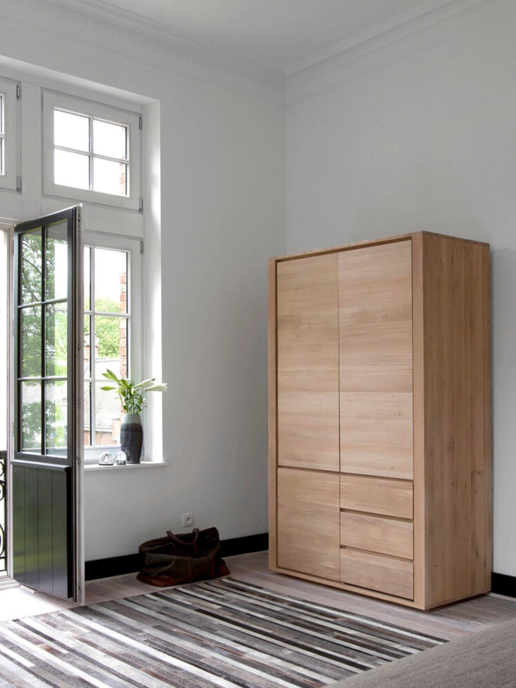 Ethnicraft Shadow Dresser in light oak, positioned in a bright, modern room with large windows, showcasing its elegant and functional design as a versatile storage solution.