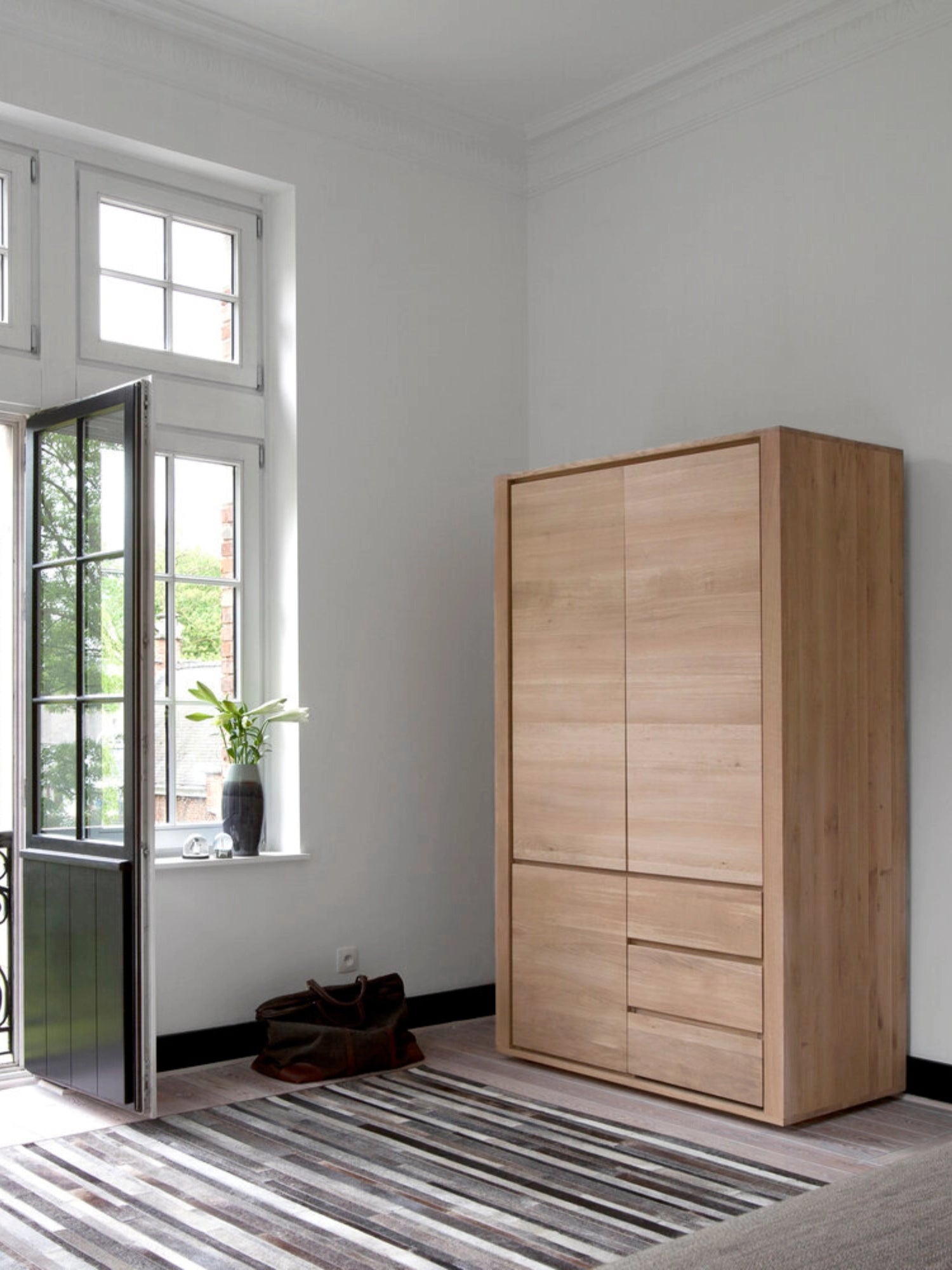 Ethnicraft Shadow Dresser in light oak, positioned in a bright, modern room with large windows, showcasing its elegant and functional design as a versatile storage solution.