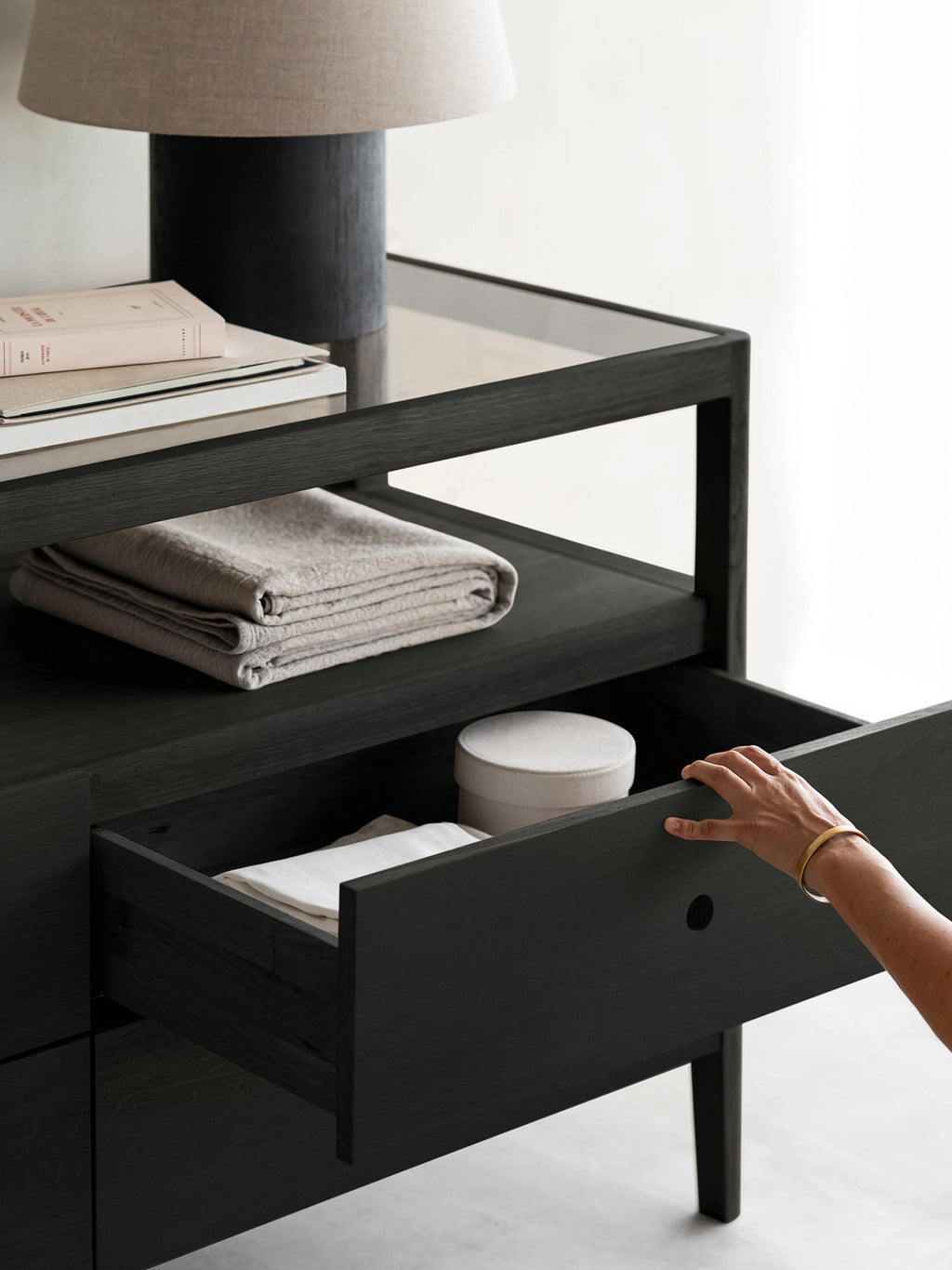 Ethnicraft Spindle Dresser in black oak, styled in a contemporary room. A hand reaches into an open drawer, revealing organized items, with a lamp and books on top.