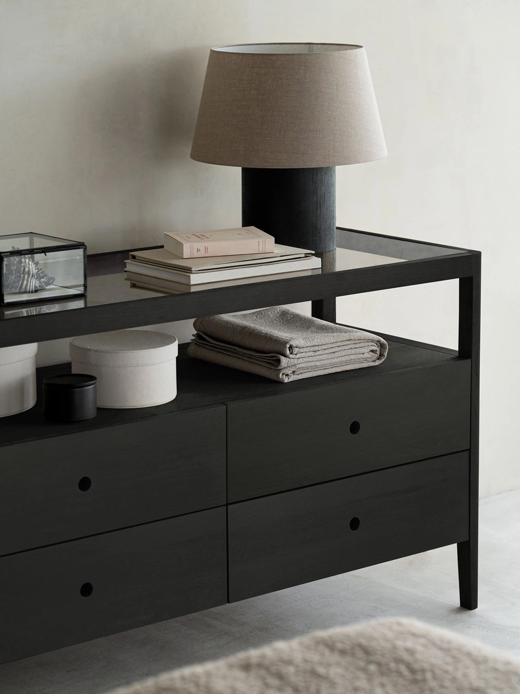 Ethnicraft Spindle Dresser in sophisticated black oak, styled in a modern living space with a lamp, books, and decorative items on its glass top and open shelf.