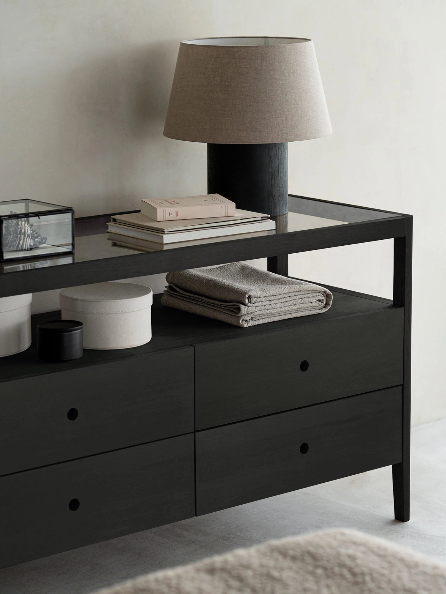 Ethnicraft Spindle Dresser in sophisticated black oak, styled in a modern living space with a lamp, books, and decorative items on its glass top and open shelf.
