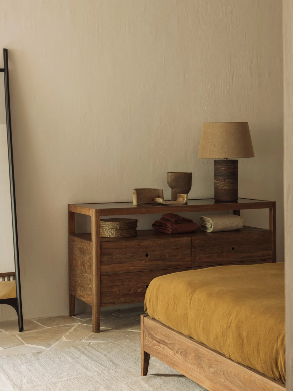 Ethnicraft Spindle Dresser in rich reclaimed teak, styled in a warm bedroom setting with a mustard yellow bedspread, a tall mirror, and a textured lamp, exuding cozy elegance.