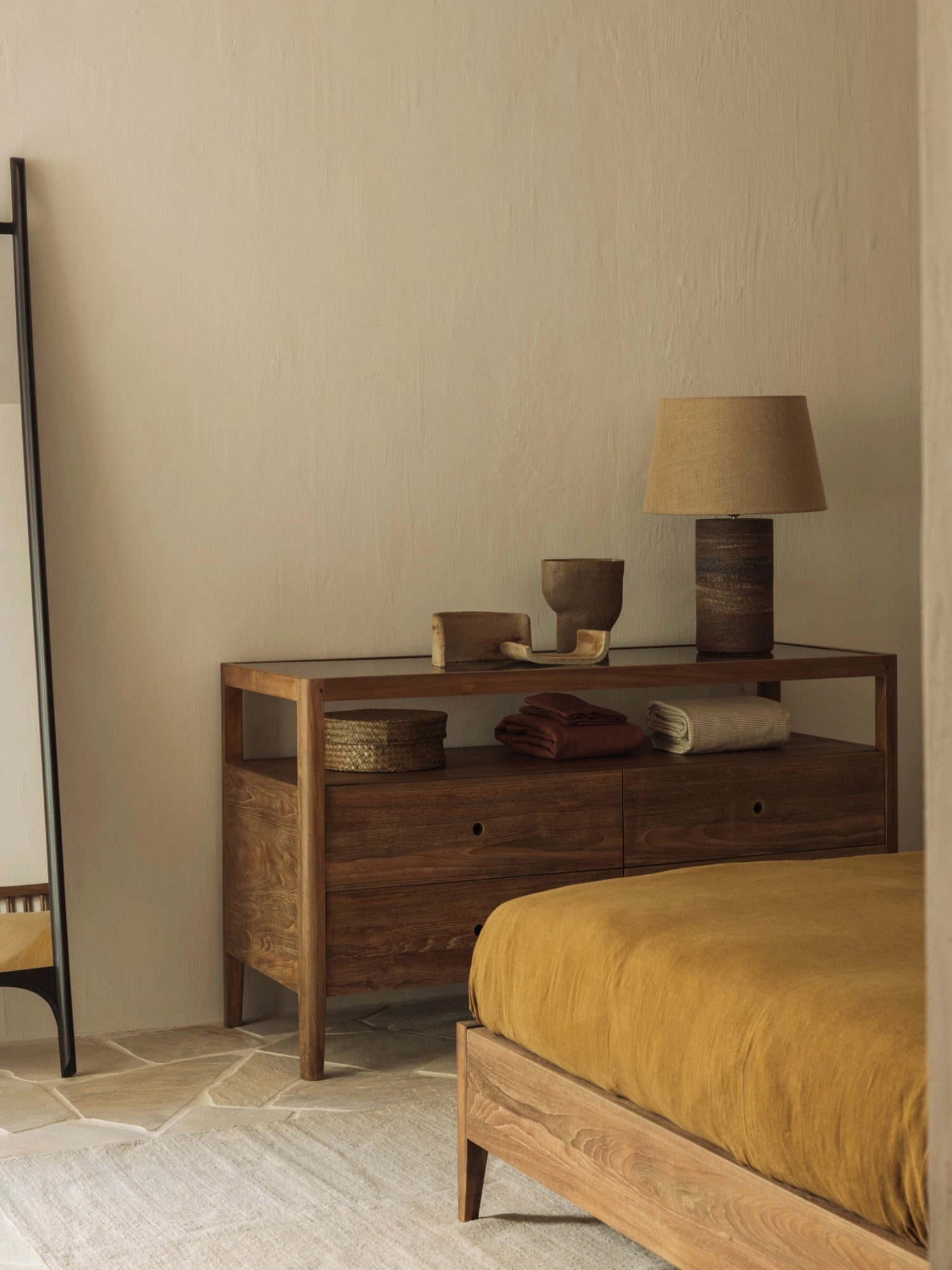 Ethnicraft Spindle Dresser in rich reclaimed teak, styled in a warm bedroom setting with a mustard yellow bedspread, a tall mirror, and a textured lamp, exuding cozy elegance.