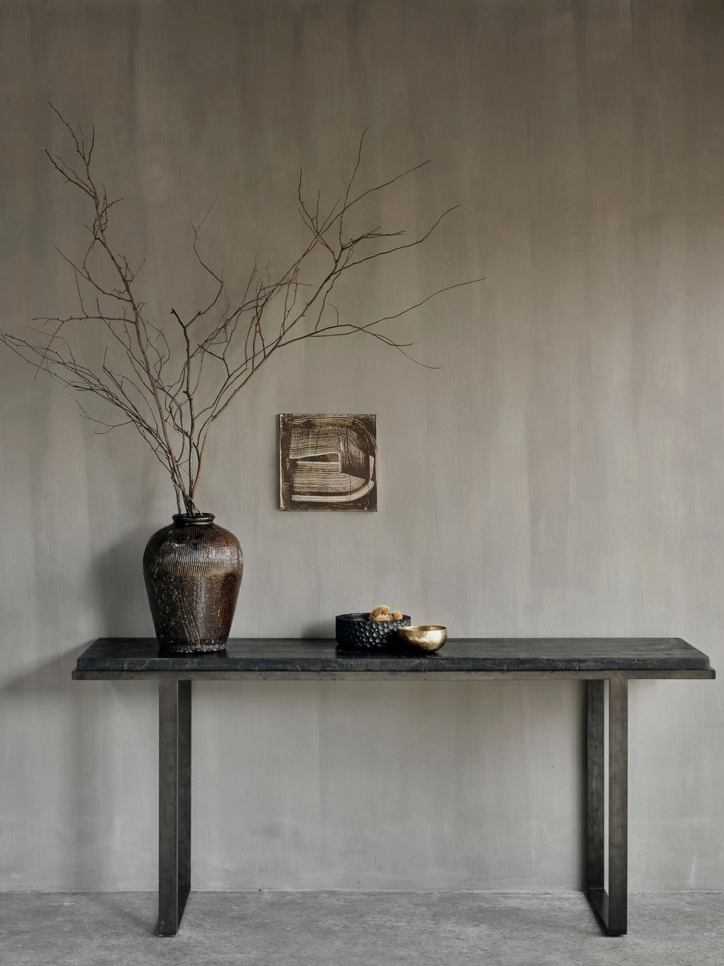 Ethnicraft Stability Console, beautifully styled in a contemporary room with a textured gray wall. The dark brown console, with its elegant textured top and metal legs, holds a large vase with branches and decorative bowls.