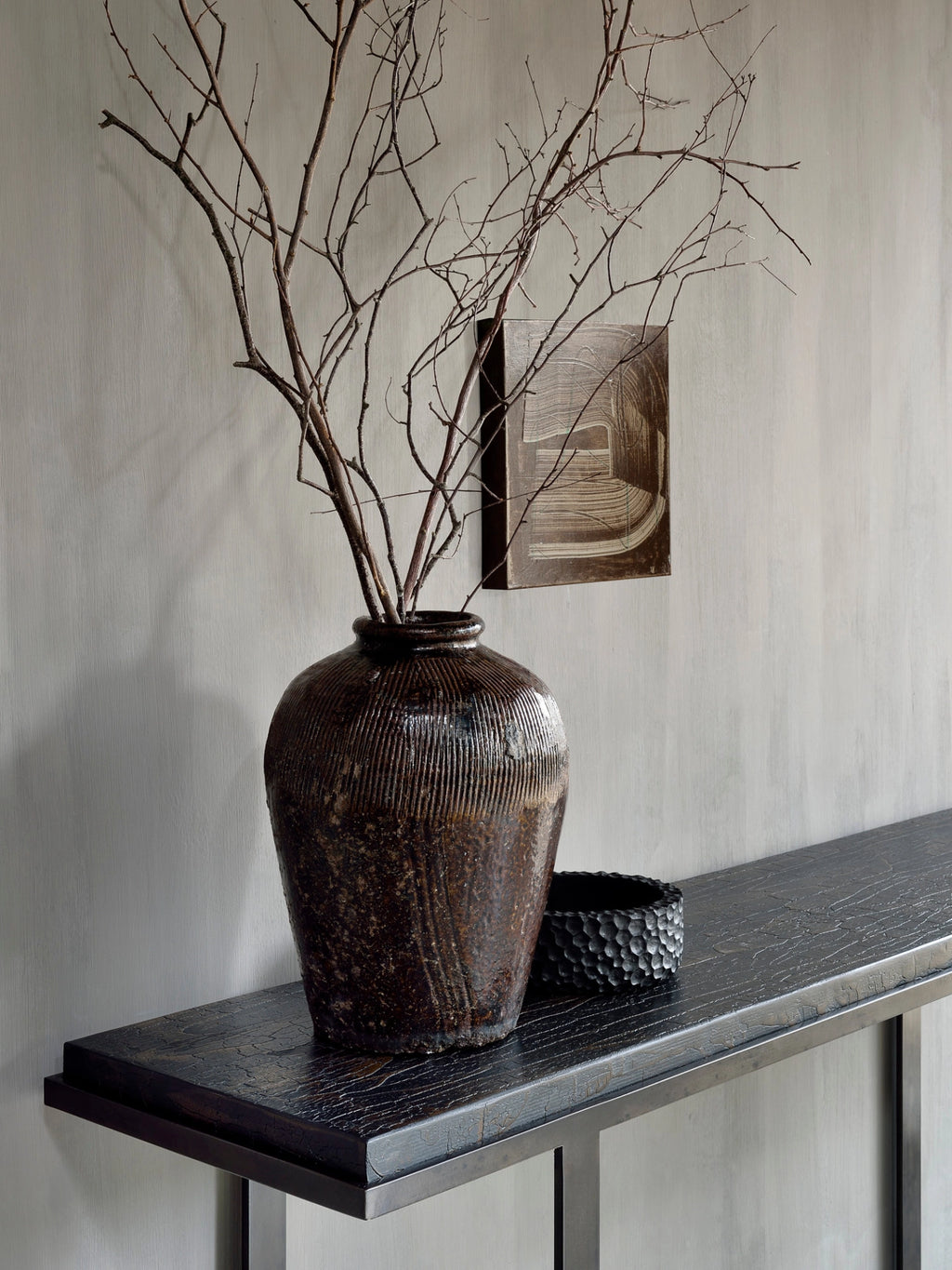 Ethnicraft Stability Console, showcasing its dark brown textured top and sleek metal frame, styled in a modern interior. A large, rustic vase filled with branches and a decorative bowl adorn the elegant console, enhancing the room's aesthetic.