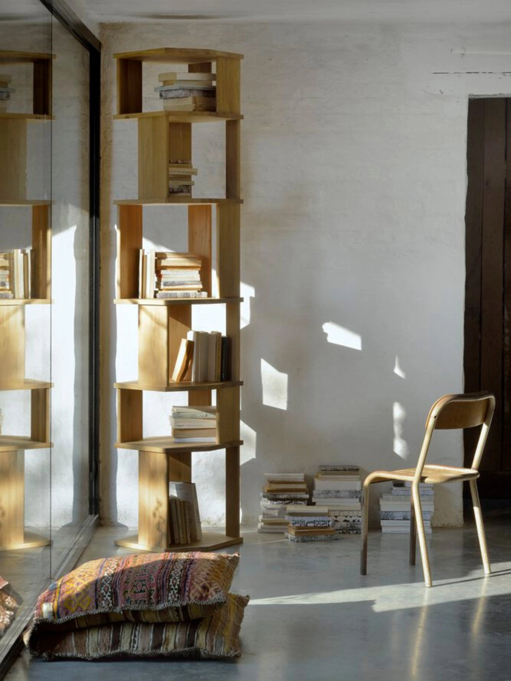 Ethnicraft Stairs Column in light natural oak, a tall, elegant modular bookshelf with open compartments, filled with books in a sunlit room with a wooden chair and patterned cushions.