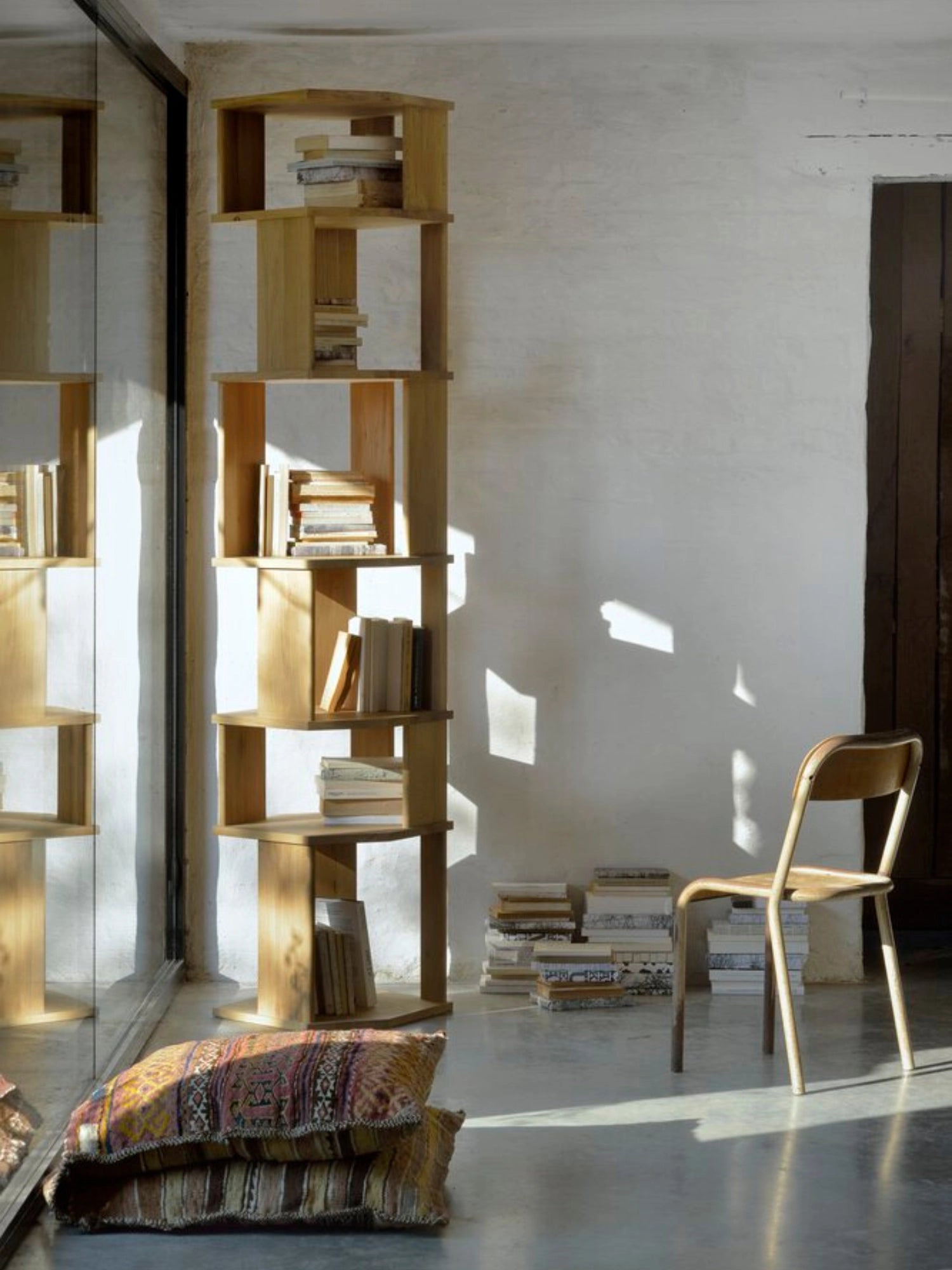 Ethnicraft Stairs Column in light natural oak, a tall, elegant modular bookshelf with open compartments, filled with books in a sunlit room with a wooden chair and patterned cushions.