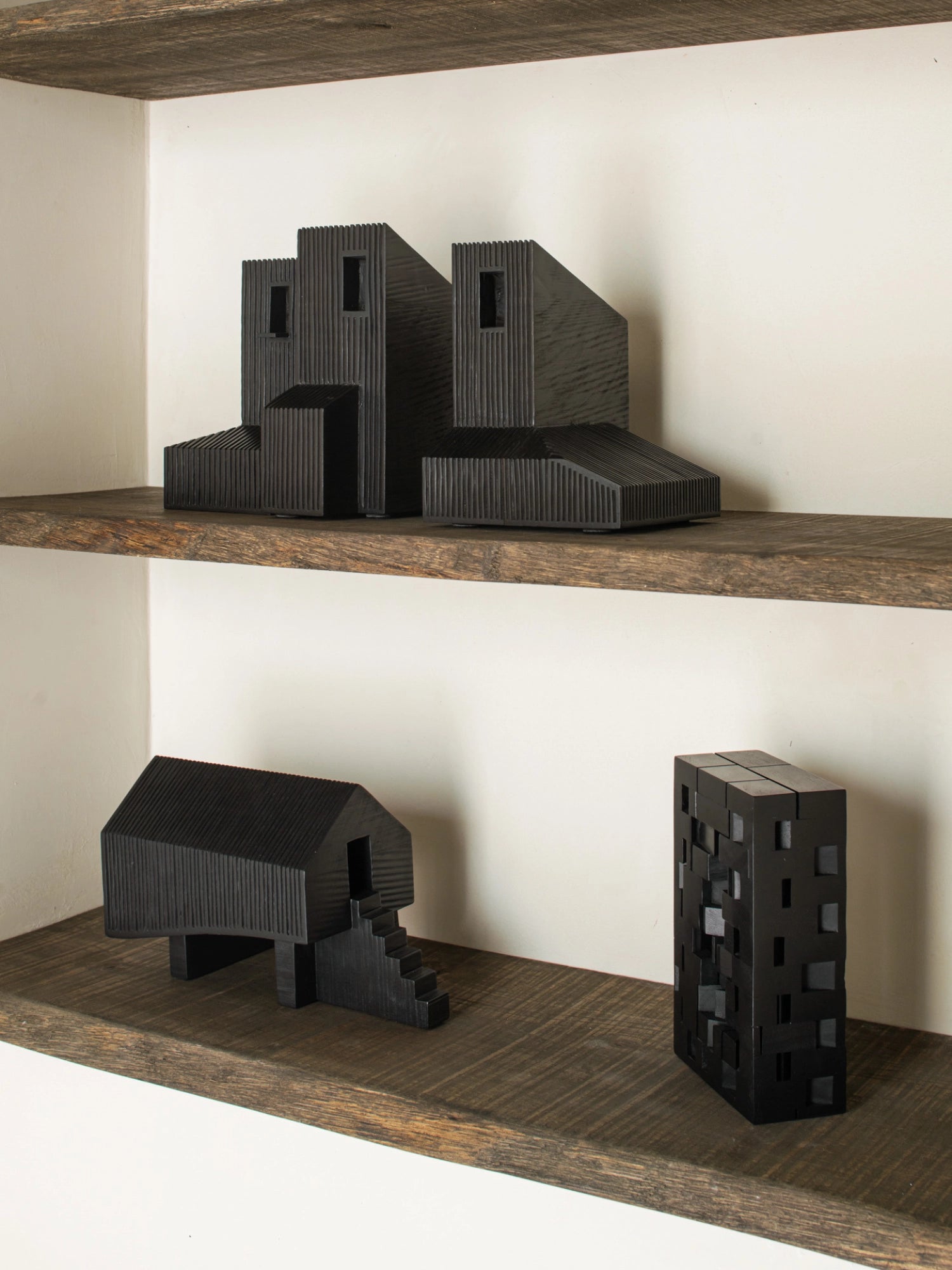 Ethnicraft Stilt House Object. A collection of three dark black wooden house sculptures with varying designs and ribbed textures, elegantly arranged on rustic wooden shelves, creating a sophisticated and artistic display.