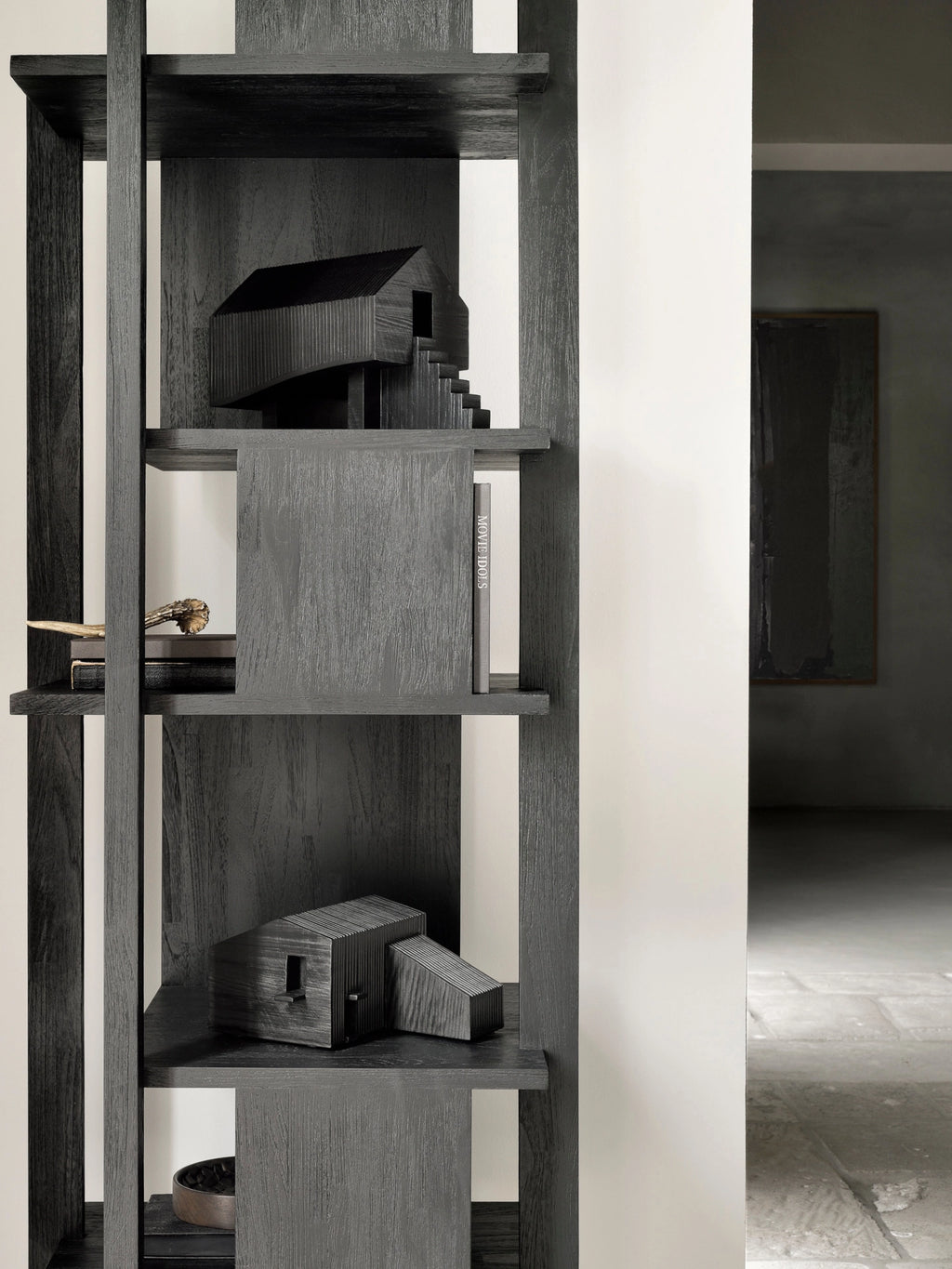 Ethnicraft Stilt House Object. A modern, dark stained wooden decorative house object with a ribbed texture and stairs, displayed on a contemporary black wooden bookshelf, adding an artistic touch to the room.