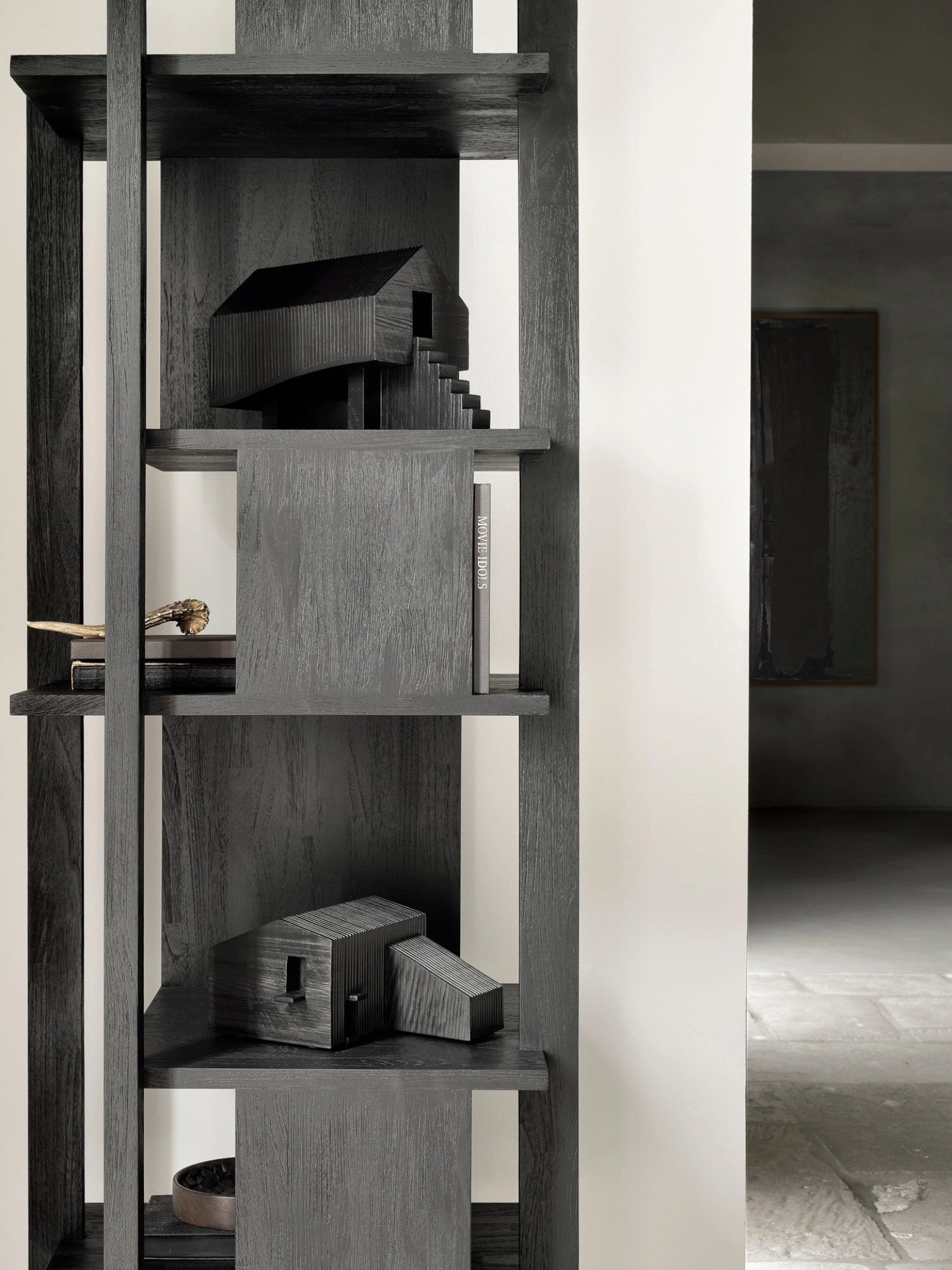 Ethnicraft Stilt House Object. A modern, dark stained wooden decorative house object with a ribbed texture and stairs, displayed on a contemporary black wooden bookshelf, adding an artistic touch to the room.