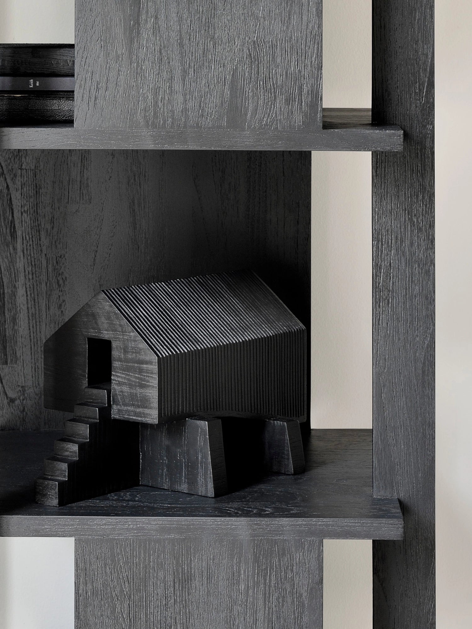 Ethnicraft Stilt House Object. A single dark black wooden house sculpture with a ribbed roof and stairs, artfully placed on a dark wooden shelf within a modern, minimalist shelving unit, enhancing the room's aesthetic.