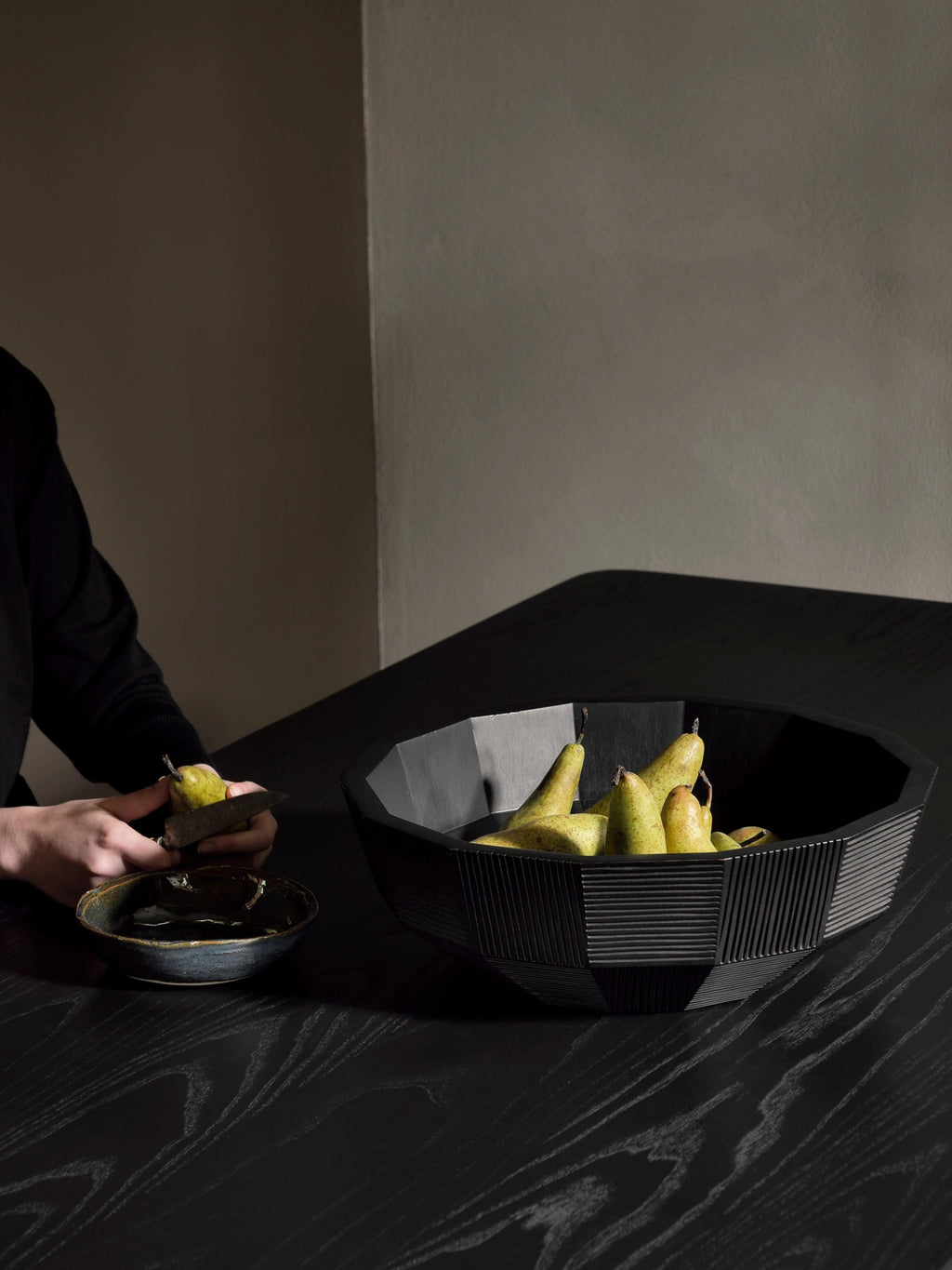 Ethnicraft Striped Bowl, a large, elegant black wooden bowl with intricate geometric striped patterns, filled with fresh green pears, on a dark wooden table in a stylish home setting.