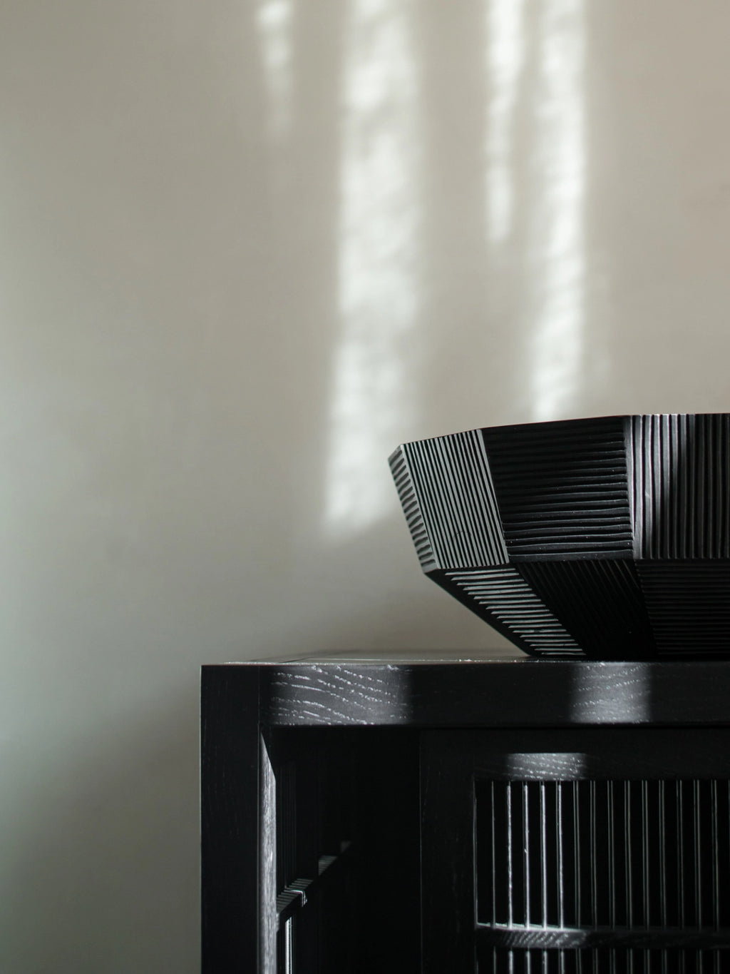 Ethnicraft Striped Bowl, a striking black wooden bowl with textured, linear striped patterns, partially visible on a dark wooden cabinet, illuminated by natural light creating shadows.