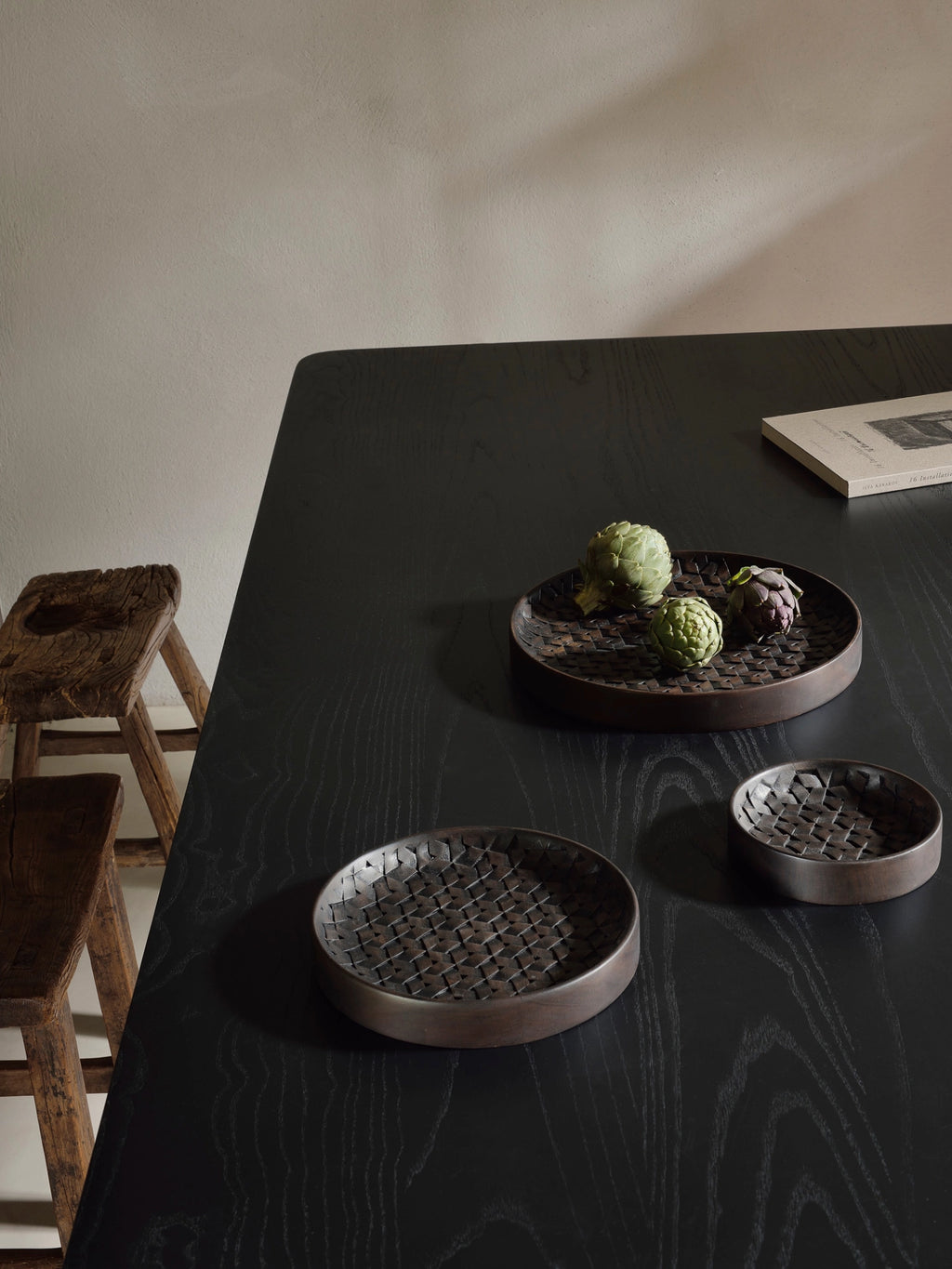 Ethnicraft Tampa Boards Set, three dark brown wooden serving boards with intricate woven patterns, holding fresh artichokes, elegantly arranged on a sleek black wooden table in a contemporary setting.