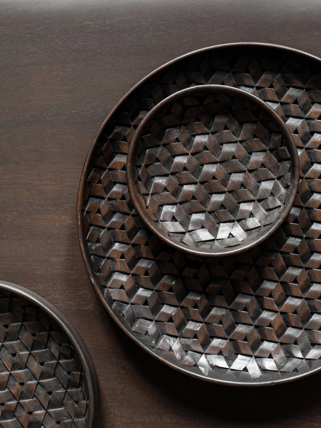 Ethnicraft Tampa Boards Set, an overhead view of three round dark brown wooden serving boards, featuring a detailed geometric woven pattern, artfully arranged on a dark wooden surface.