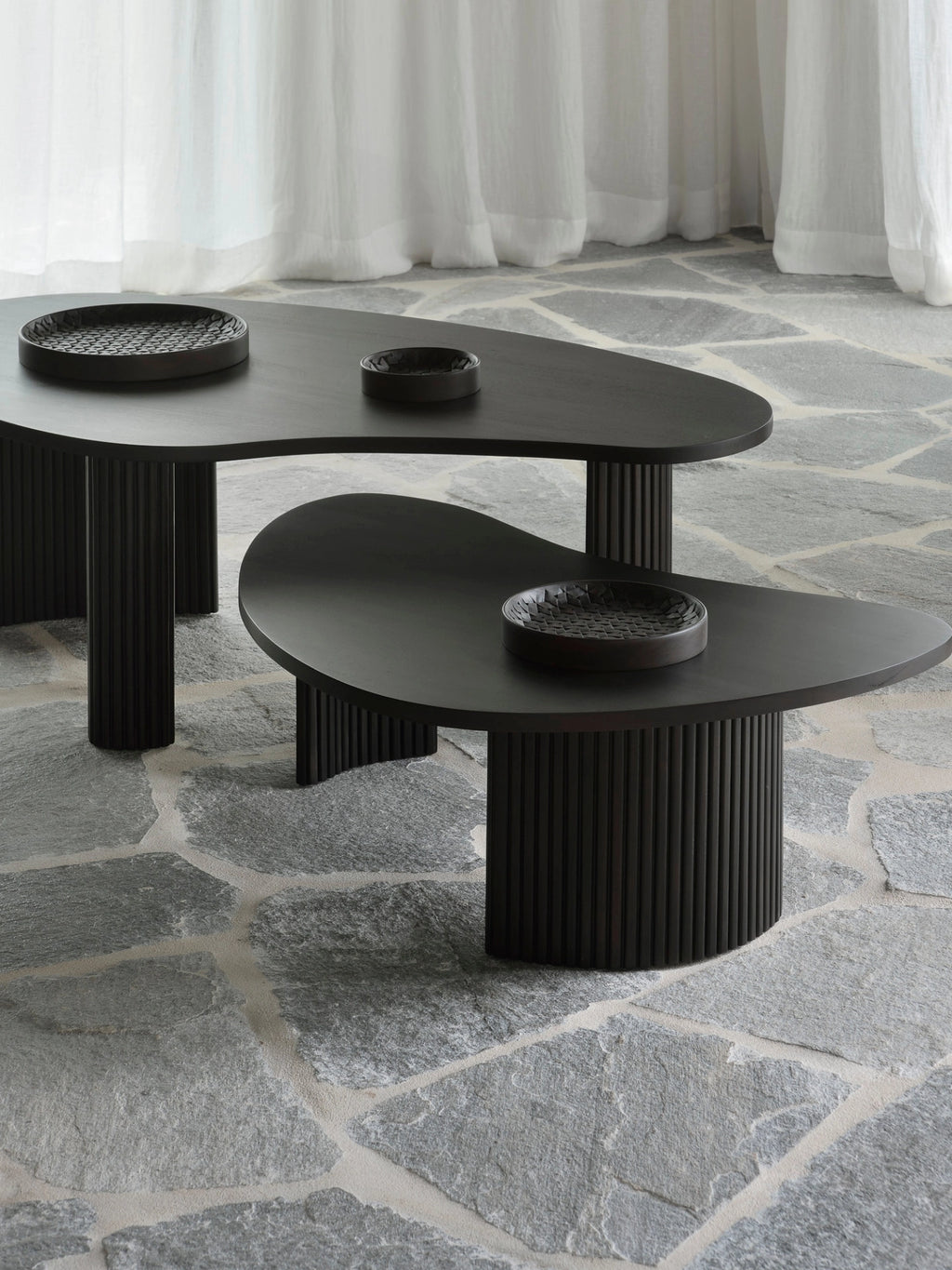 Ethnicraft Tampa Boards Set, two dark brown wooden serving boards with geometric woven patterns, resting on modern black coffee tables with fluted bases, set against a stylish stone floor and sheer curtains.