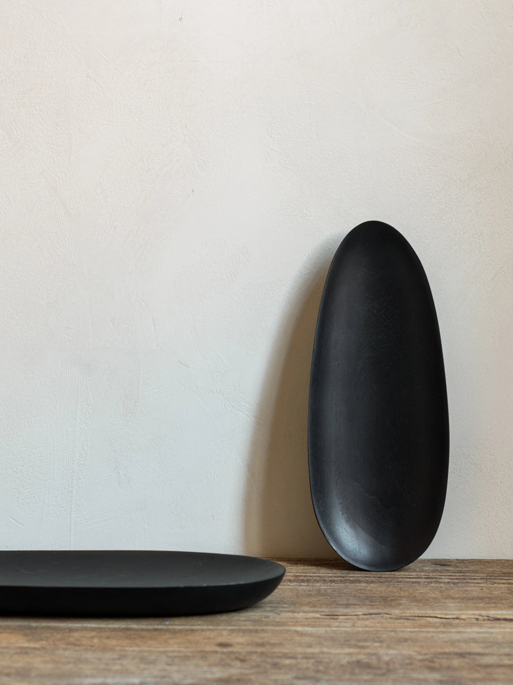 Ethnicraft Thin Oval Boards Set in mahogany black, with one large oval board standing upright and another lying flat, showcasing their elegant, minimalist design against a textured light-colored wall and a natural wooden surface.