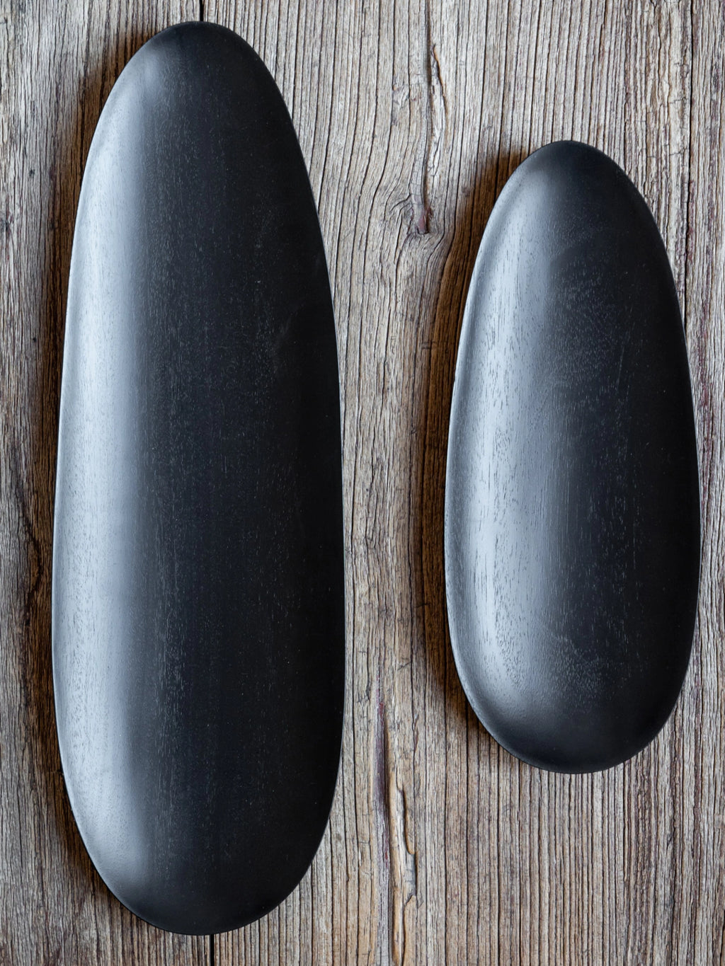 Ethnicraft Thin Oval Boards Set in mahogany black, featuring two distinctively sized, smooth, dark oval boards with a subtle wood grain texture, presented on a rustic, weathered wooden surface, emphasizing their natural material.