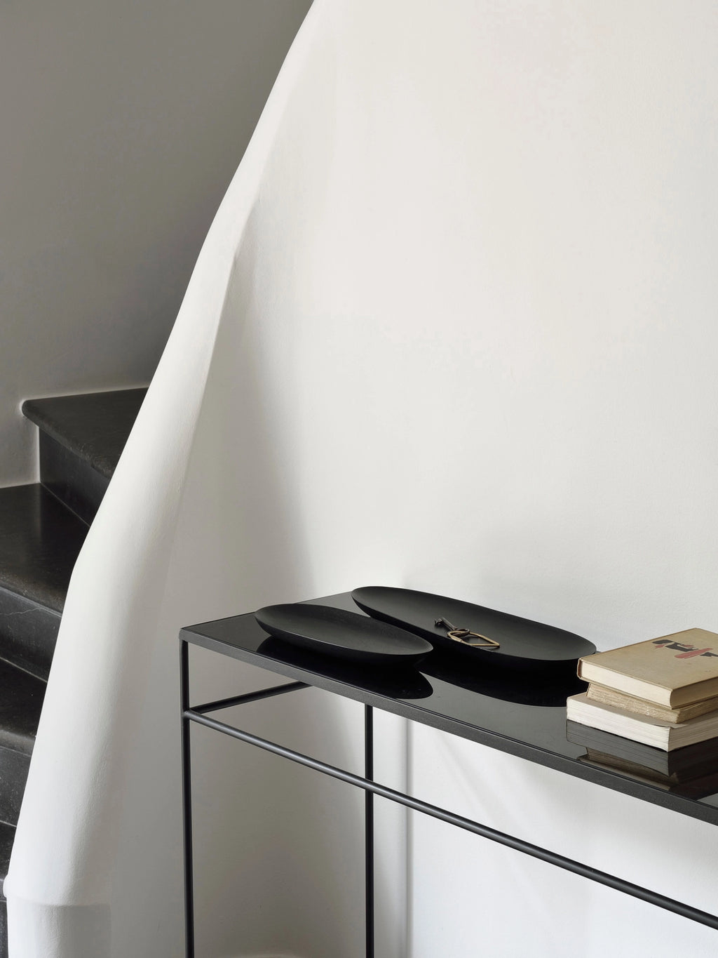 Ethnicraft Thin Oval Boards Set in mahogany black, elegantly arranged on a modern black console table with books and a key, set against a minimalist white wall and a dark staircase, highlighting contemporary home decor.
