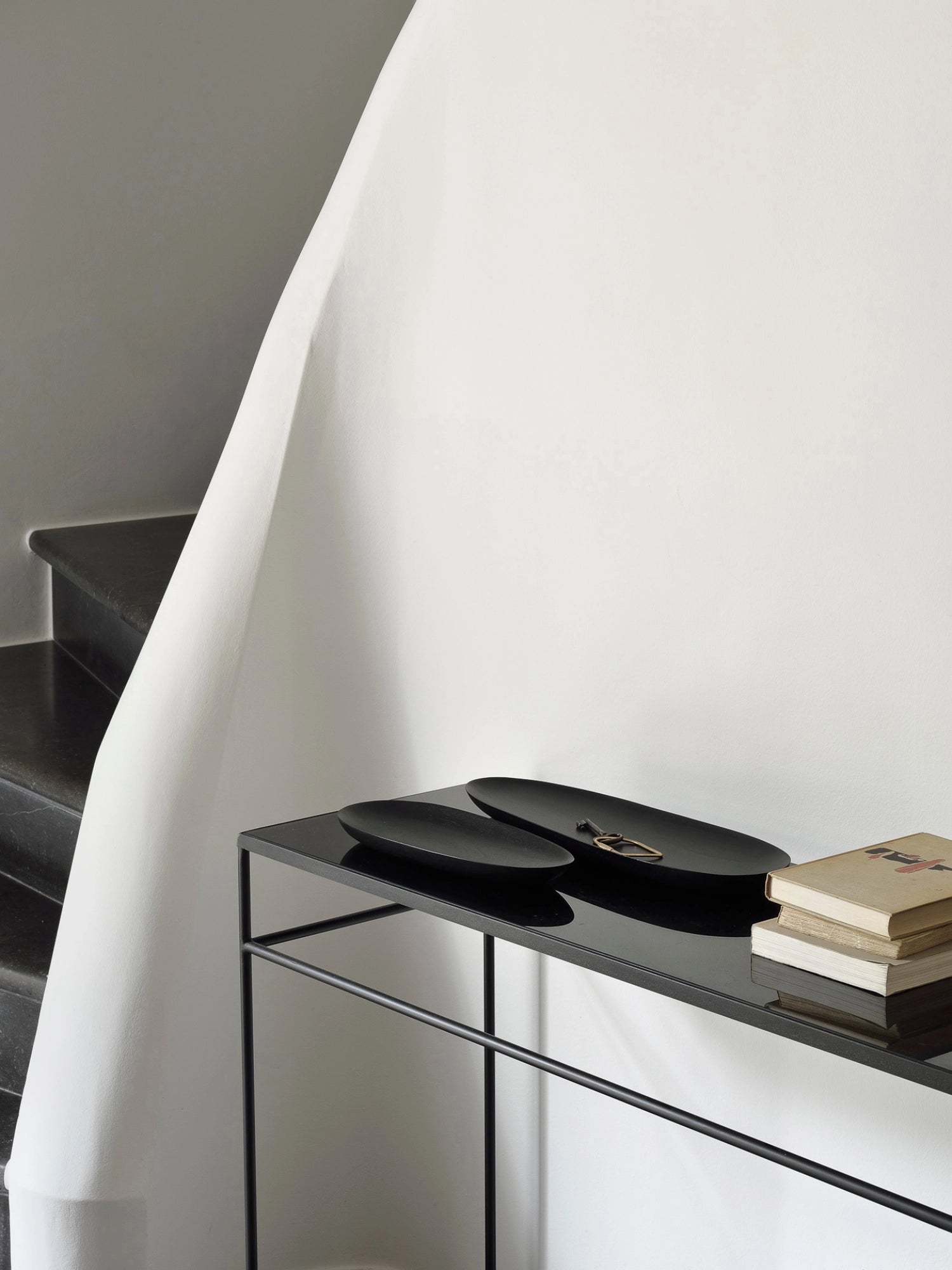Ethnicraft Thin Oval Boards Set in mahogany black, elegantly arranged on a modern black console table with books and a key, set against a minimalist white wall and a dark staircase, highlighting contemporary home decor.