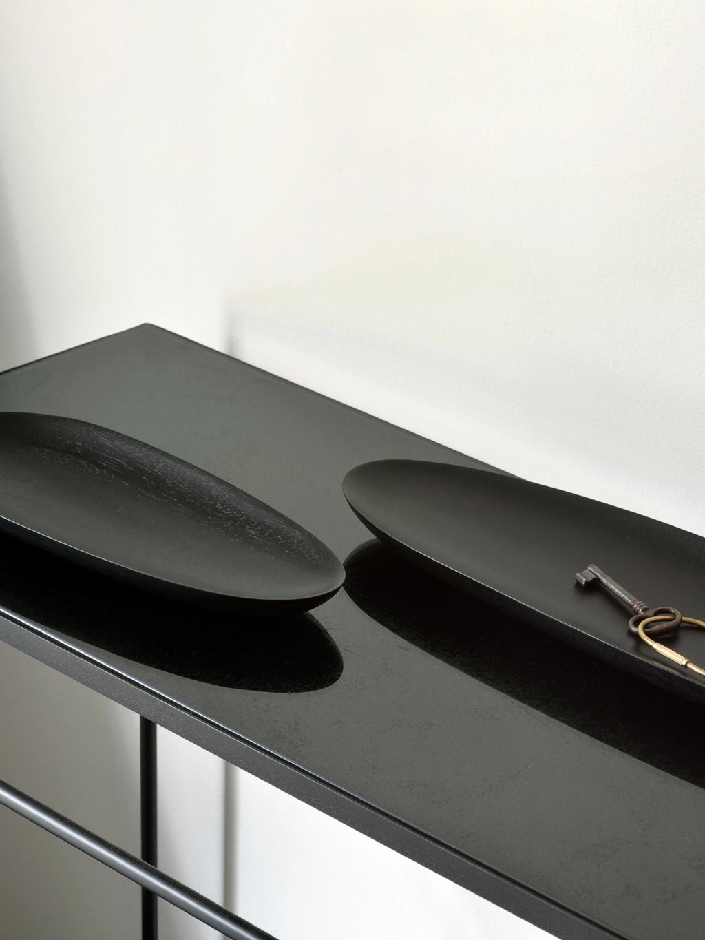 Ethnicraft Thin Oval Boards Set in mahogany black, showcasing two sleek, dark oval boards on a glossy black console table, with a vintage key set resting on one board, against a clean white wall.