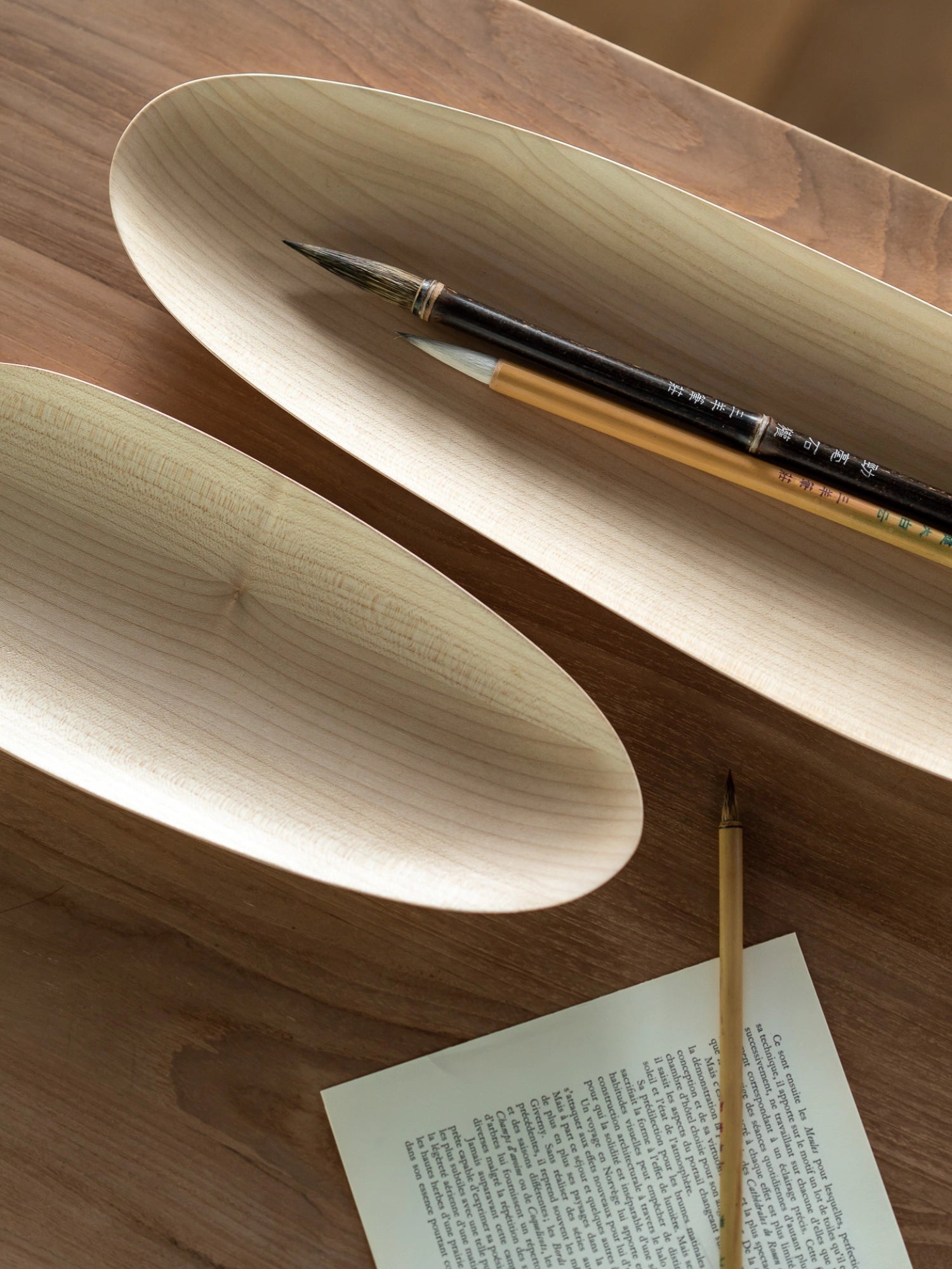 Ethnicraft Thin Oval Boards Set in sycamore, featuring two light-colored, natural wood oval boards with visible grain, accompanied by traditional calligraphy brushes and paper, displayed on a warm wooden table, exuding a serene aesthetic.