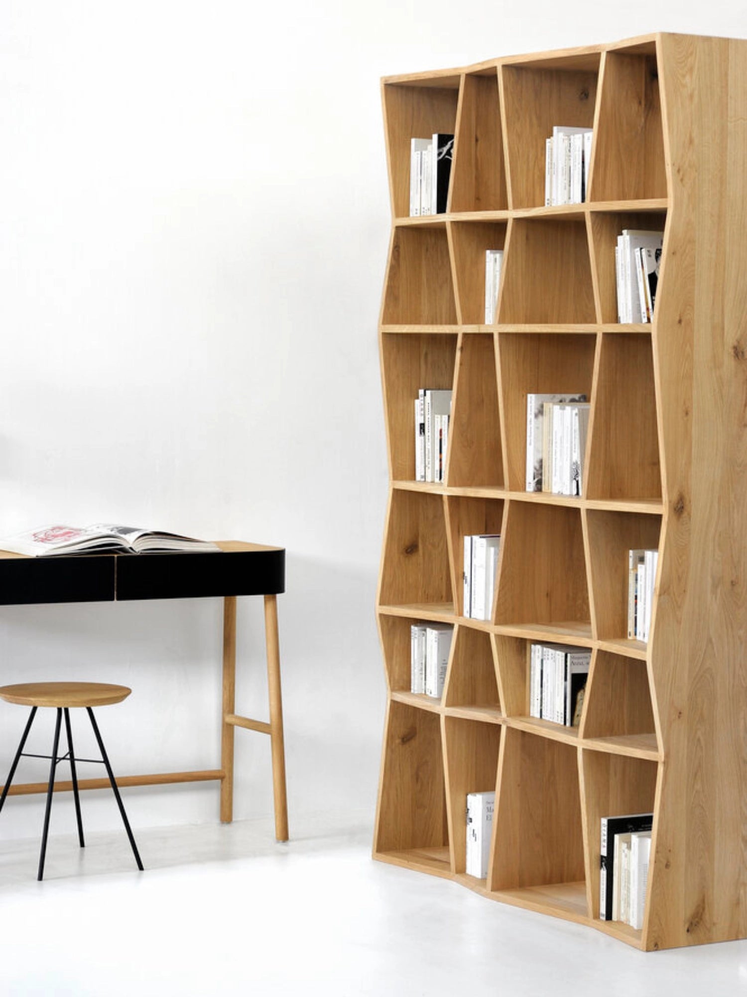 Ethnicraft Z Rack, a tall, modern light oak wood shelving unit with unique Z-shaped compartments, filled with books, positioned next to a minimalist desk in a bright room.