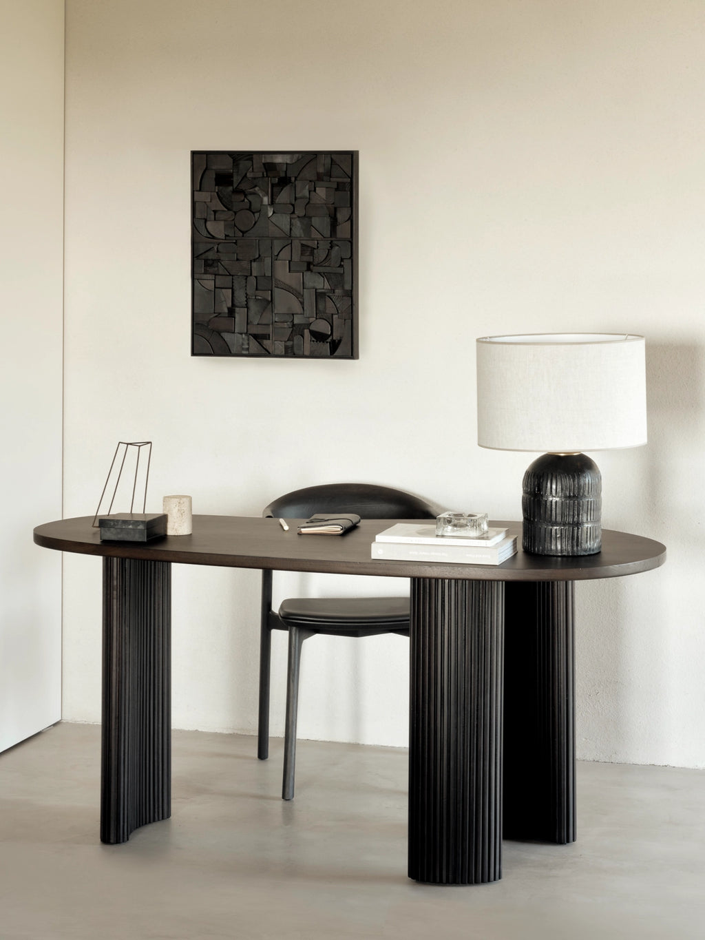 Ethnicraft Boomerang Desk in dark stained oak, showcasing its distinctive oval top and fluted cylindrical legs, accompanied by a black chair and a stylish lamp in a minimalist room.