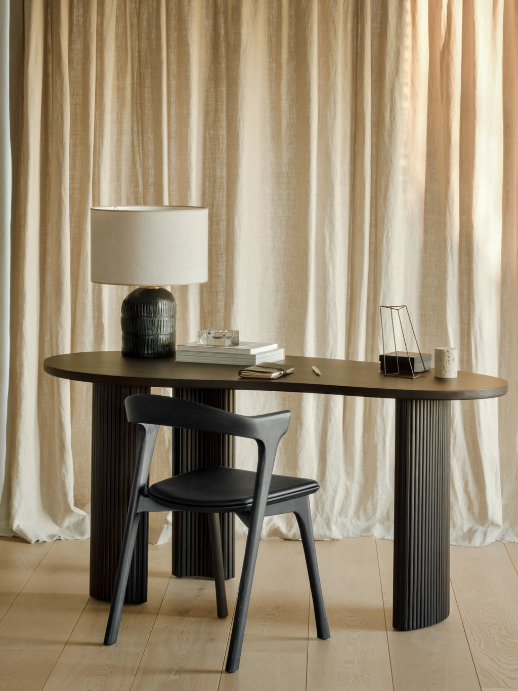 Ethnicraft Boomerang Desk in dark stained oak, featuring a unique oval tabletop and fluted cylindrical legs, paired with a black chair and a modern lamp against a draped curtain background.