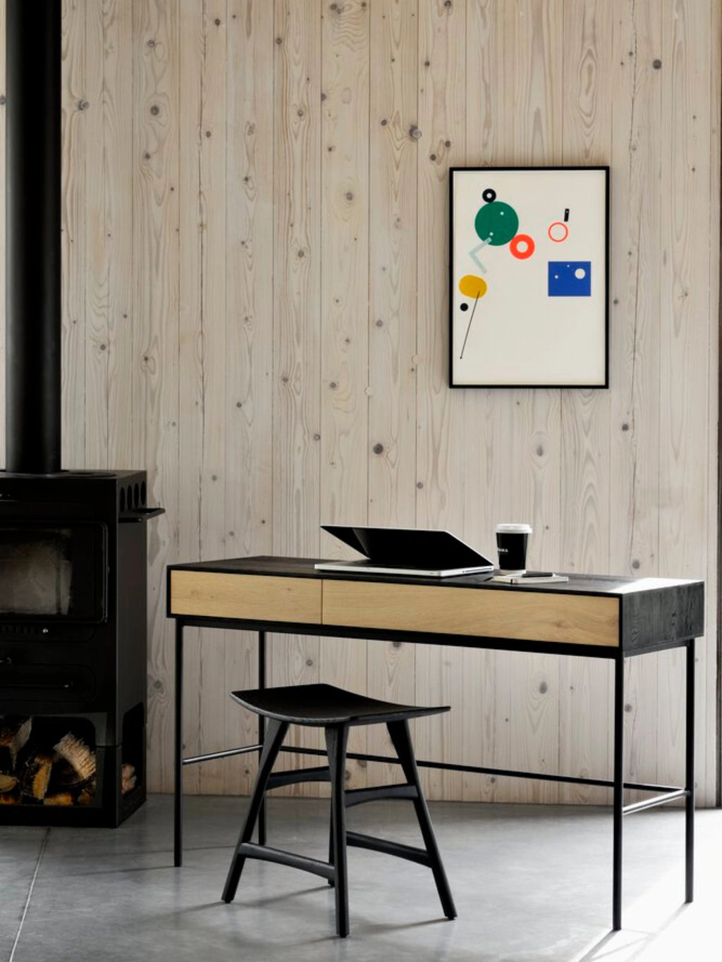 Ethnicraft Blackbird Desk in a cozy, rustic-modern room. The dark oak desk with black metal frame is set against a light wood paneled wall, featuring a laptop, coffee cup, and a stylish abstract art print above.