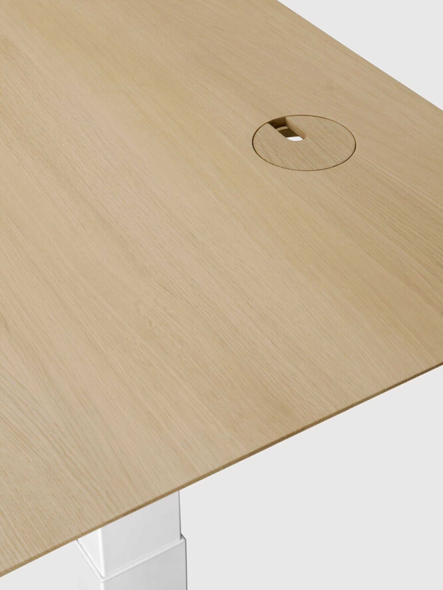 Ethnicraft Bok Adjustable Desk, a close-up detail of the light oak wood tabletop, revealing its smooth, natural grain and a discreet circular cable management grommet. This elegant, functional design enhances any modern office.