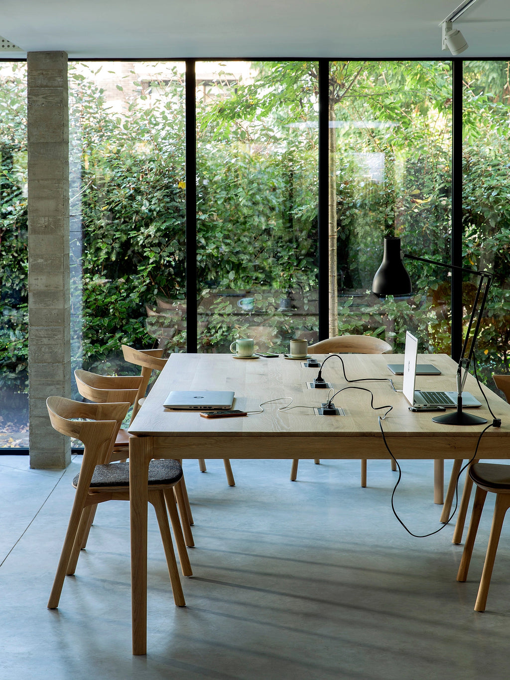 Ethnicraft Bok Cowork Desk in a modern office with large windows, showcasing the long, natural oak table with integrated power, surrounded by stylish wooden chairs.