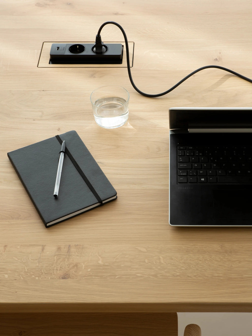 Ethnicraft Bok Cowork Desk surface detail, showing a built-in power outlet with a laptop and notebook, highlighting the desk's functional design and natural oak finish.