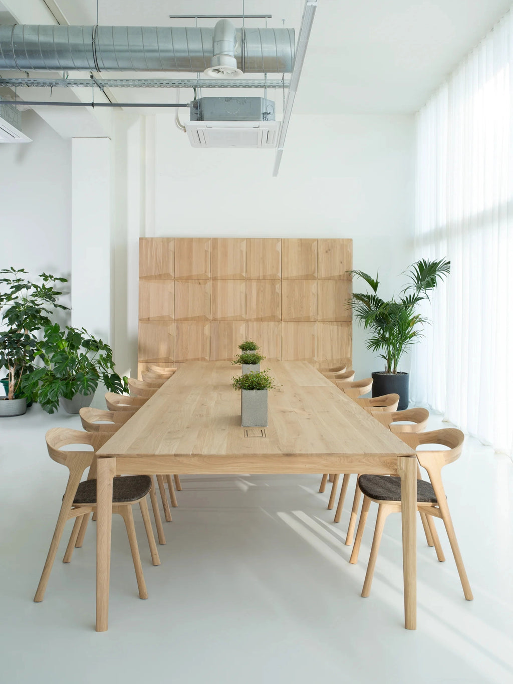 Ethnicraft Bok Cowork Desk in a spacious, bright office setting, featuring a long, natural oak table surrounded by matching wooden chairs, with integrated power outlets.