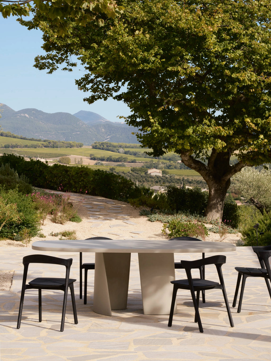 Ethnicraft Canyon Outdoor Dining Table, a contemporary oval outdoor dining table crafted from a light greige mineral composite with a smooth, raw finish and two sculptural pedestal bases. Paired with matte black solid wood outdoor dining chairs featuring curved backrests and clean lines, creating an elegant outdoor dining set. Perfect for organic modern patios, rustic Mediterranean terraces, or any stylish outdoor living space.