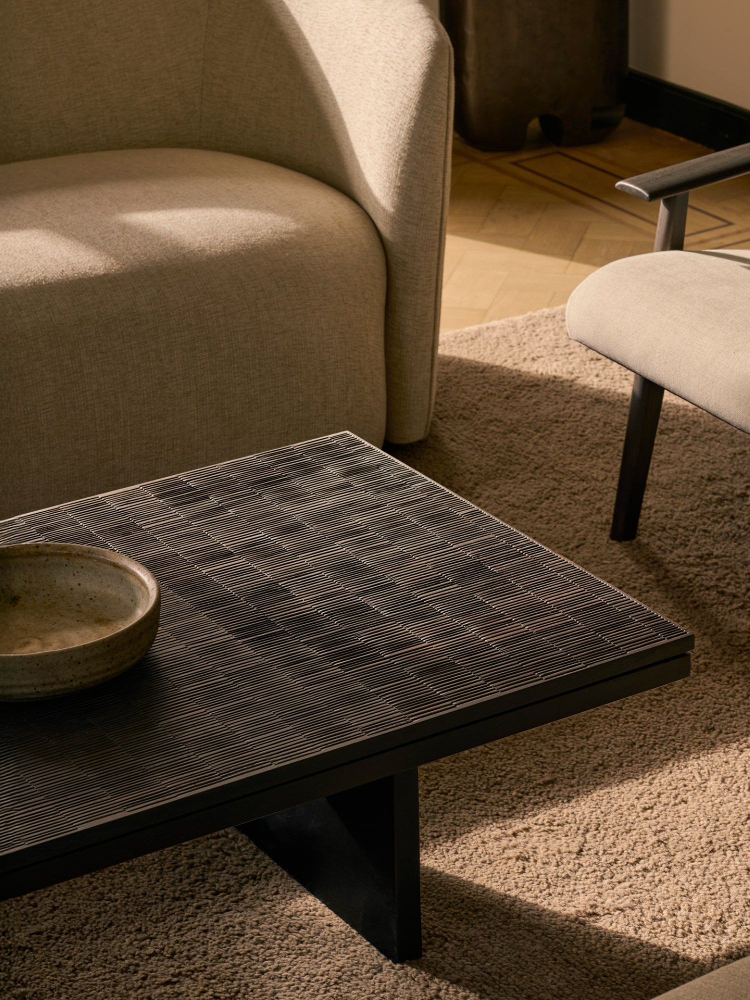 Ethnicraft Grooves Coffee Table, a close-up of the off-black teak table, revealing its distinctive grooved texture, positioned on a plush beige rug next to a modern armchair.